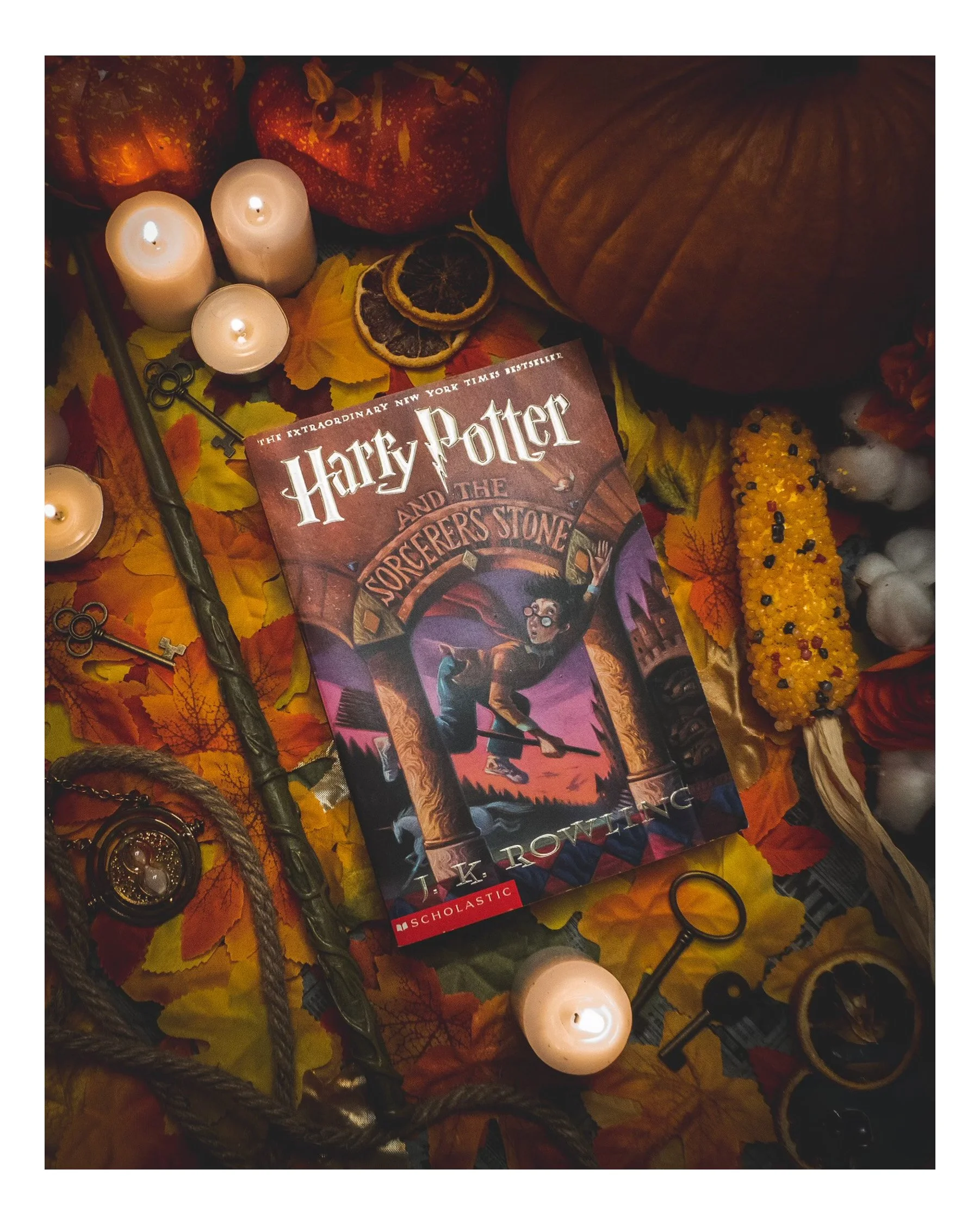A collection of autumn-themed items including orange and red pumpkins, dried orange slices, fall leaves, a Harry Potter book, candles, a key, a gray rope, and various black rings, arranged on a surface.