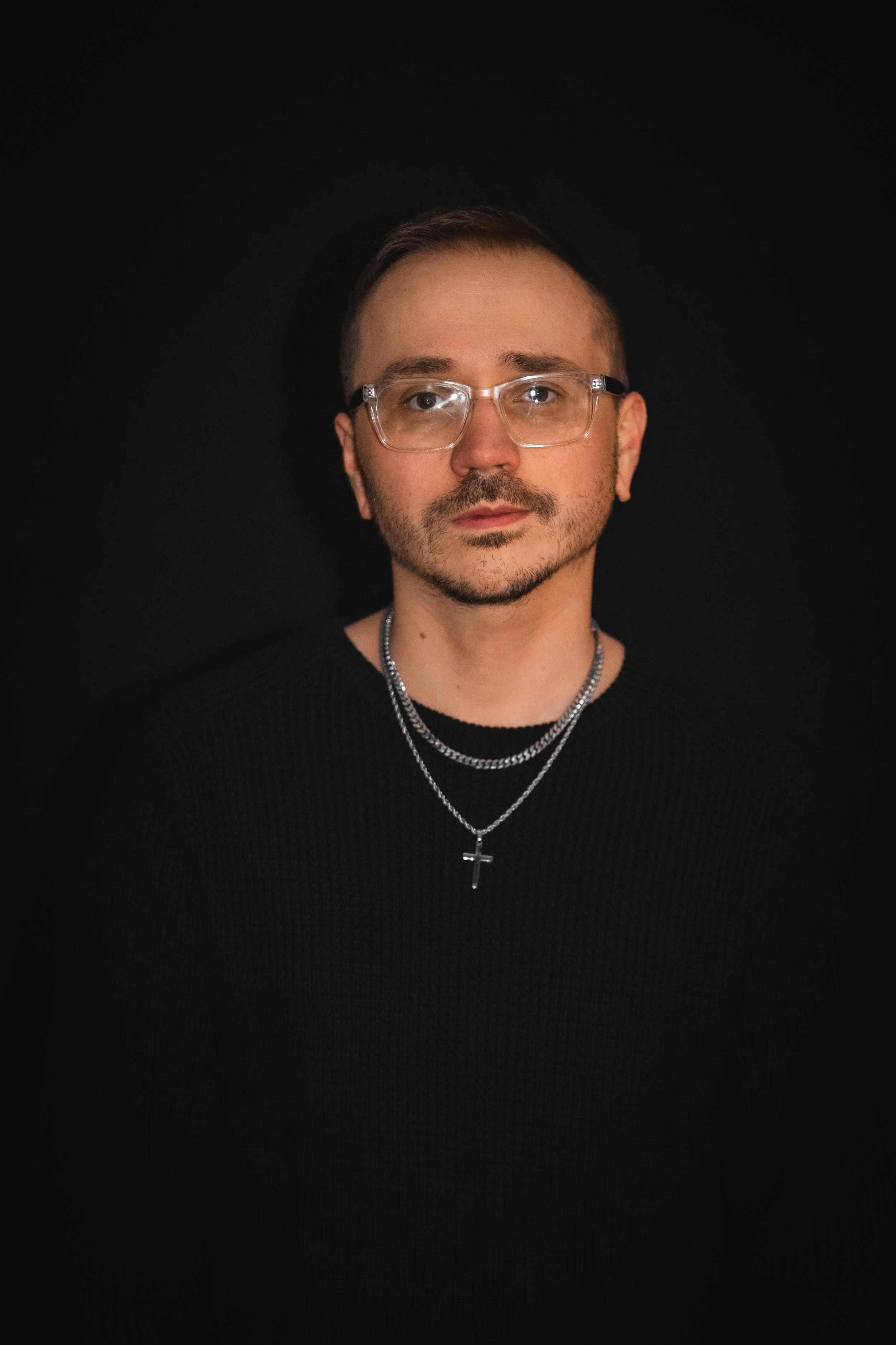 Portrait of a person with short hair, wearing glasses, a black shirt, and layered silver necklaces with a cross pendant, against a black background.