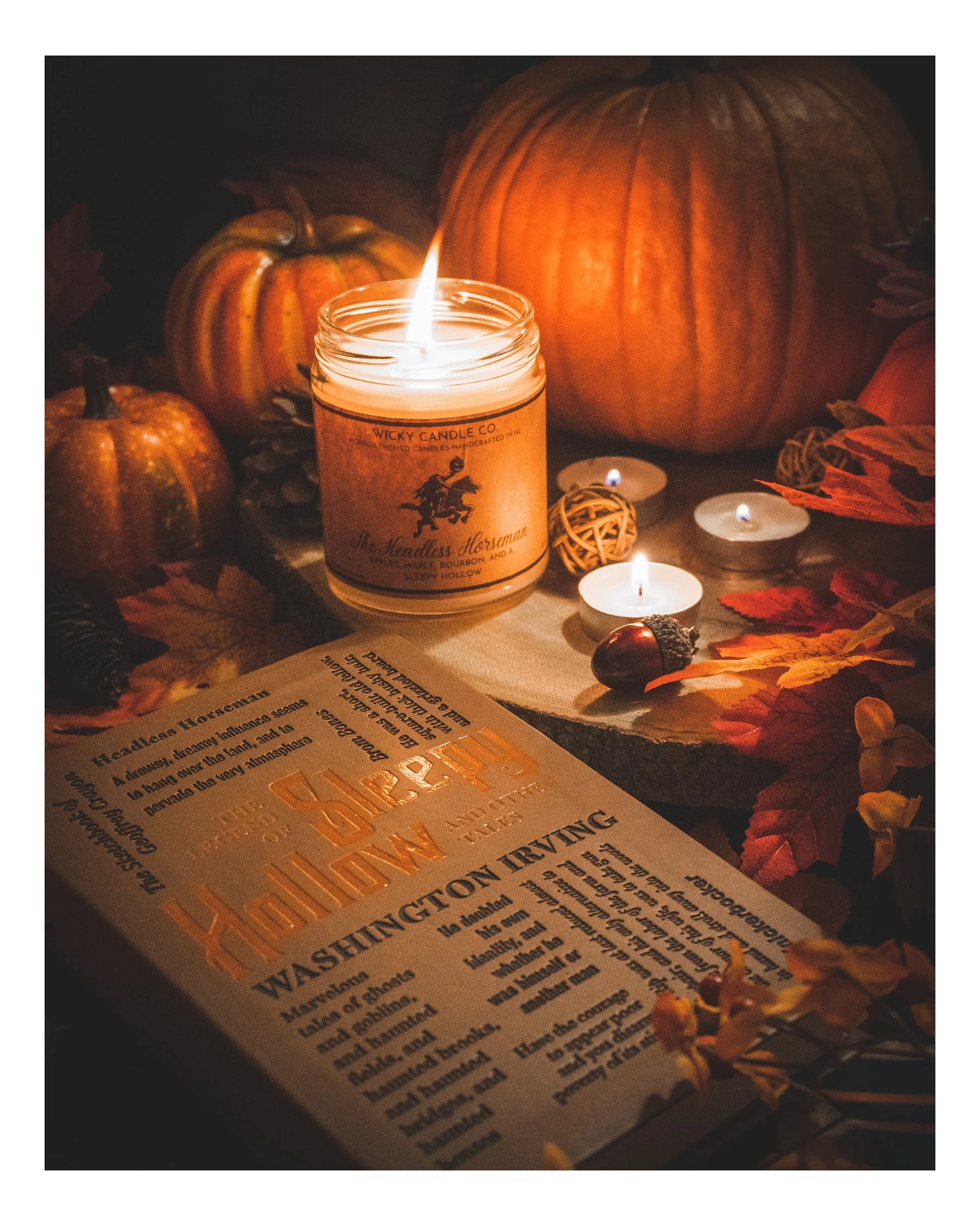 A lit candle with a label reading 'Wicky Candle Co.' surrounded by small pumpkins and autumn leaves, creating a cozy autumn or Halloween display.