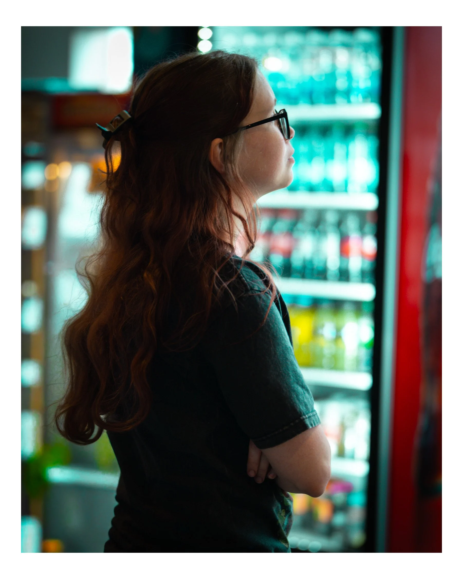 A woman with long wavy red hair, wearing glasses and a dark top, stands with arms crossed in front of a store shelf filled with colorful products, possibly beverages or snacks, in a grocery store. She is shown in profile, looking to the right.