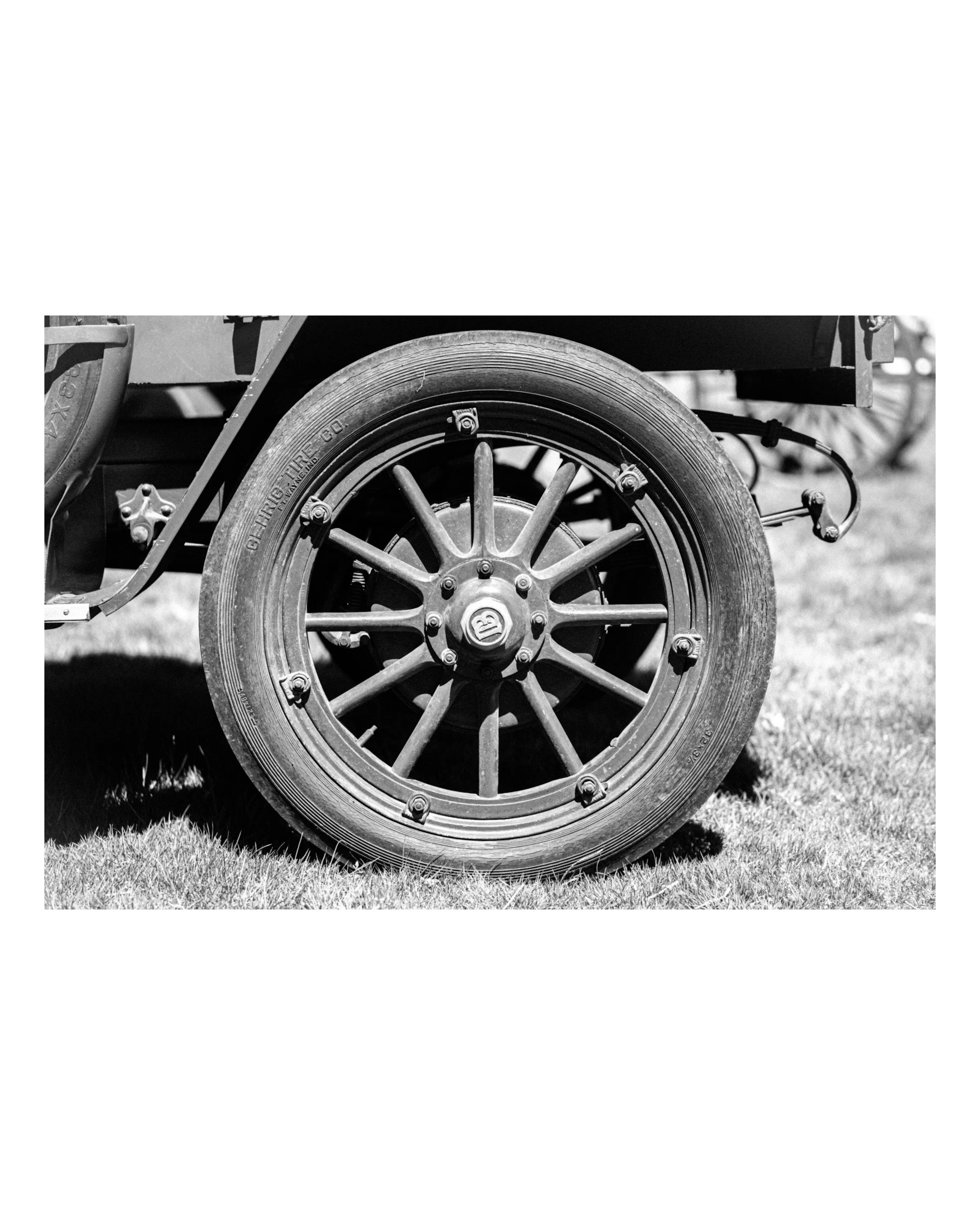 Close-up of a vintage car wheel with a detailed metal rim and tire, cast in black and white with grass underneath.