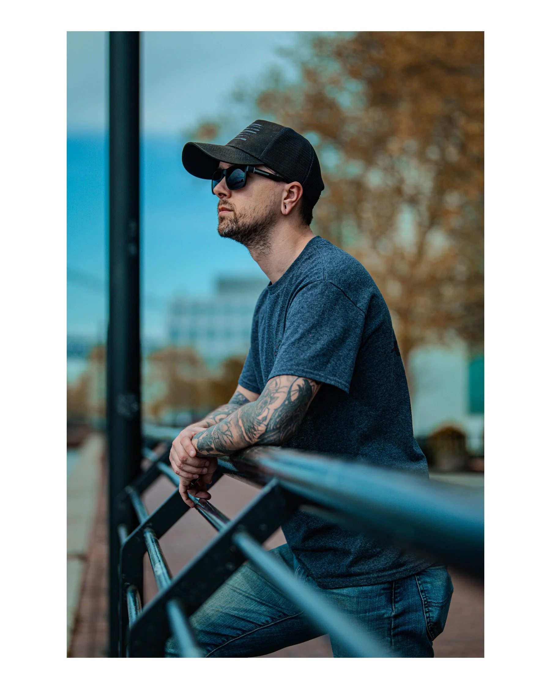 A man with tattoos on his arms wearing sunglasses, a baseball cap, a grey t-shirt, and jeans, leaning on a metal fence outdoors during autumn with blurred trees in the background.