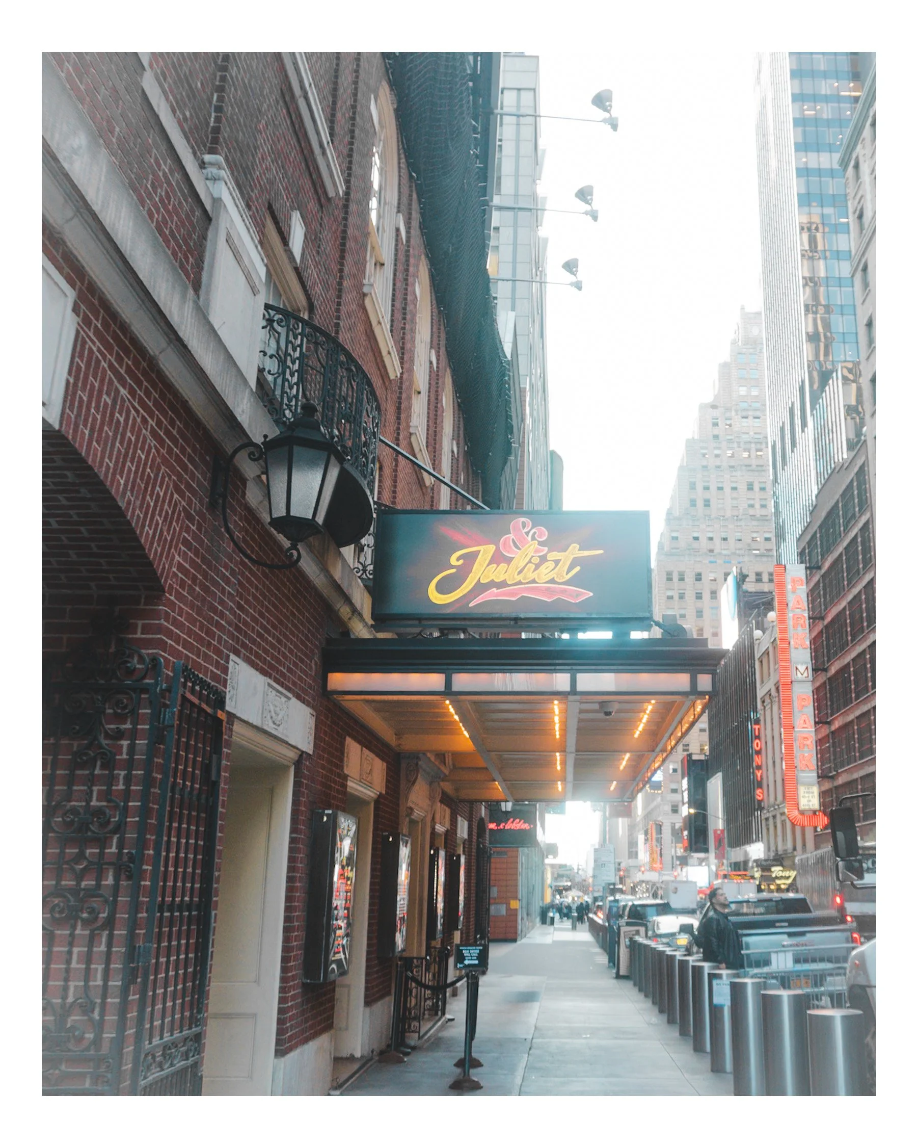 City street scene showing a brick building with above-ground windows and a black iron railing. There is a marquee with the word 'Juliet' in cursive. Along the sidewalk, there are multiple trash cans, an electronic sign near the entrance, and a few pe