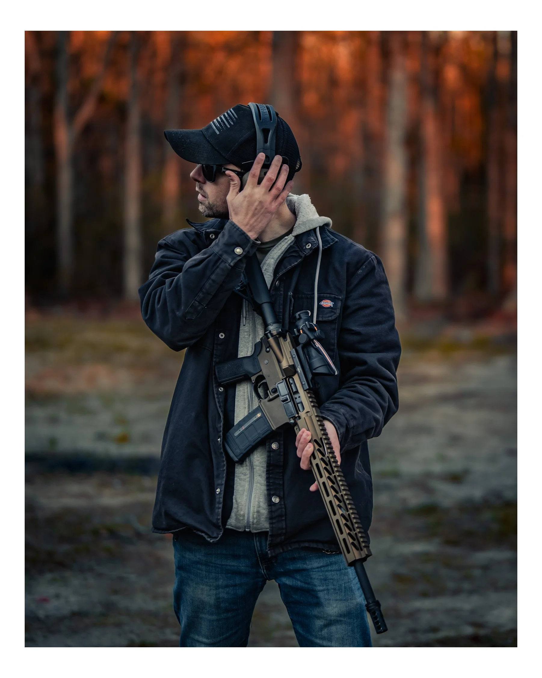 A man wearing sunglasses, a black cap, a hoodie, and a black jacket, holding a rifle with a bipod, standing outdoors during autumn with trees in the background.
