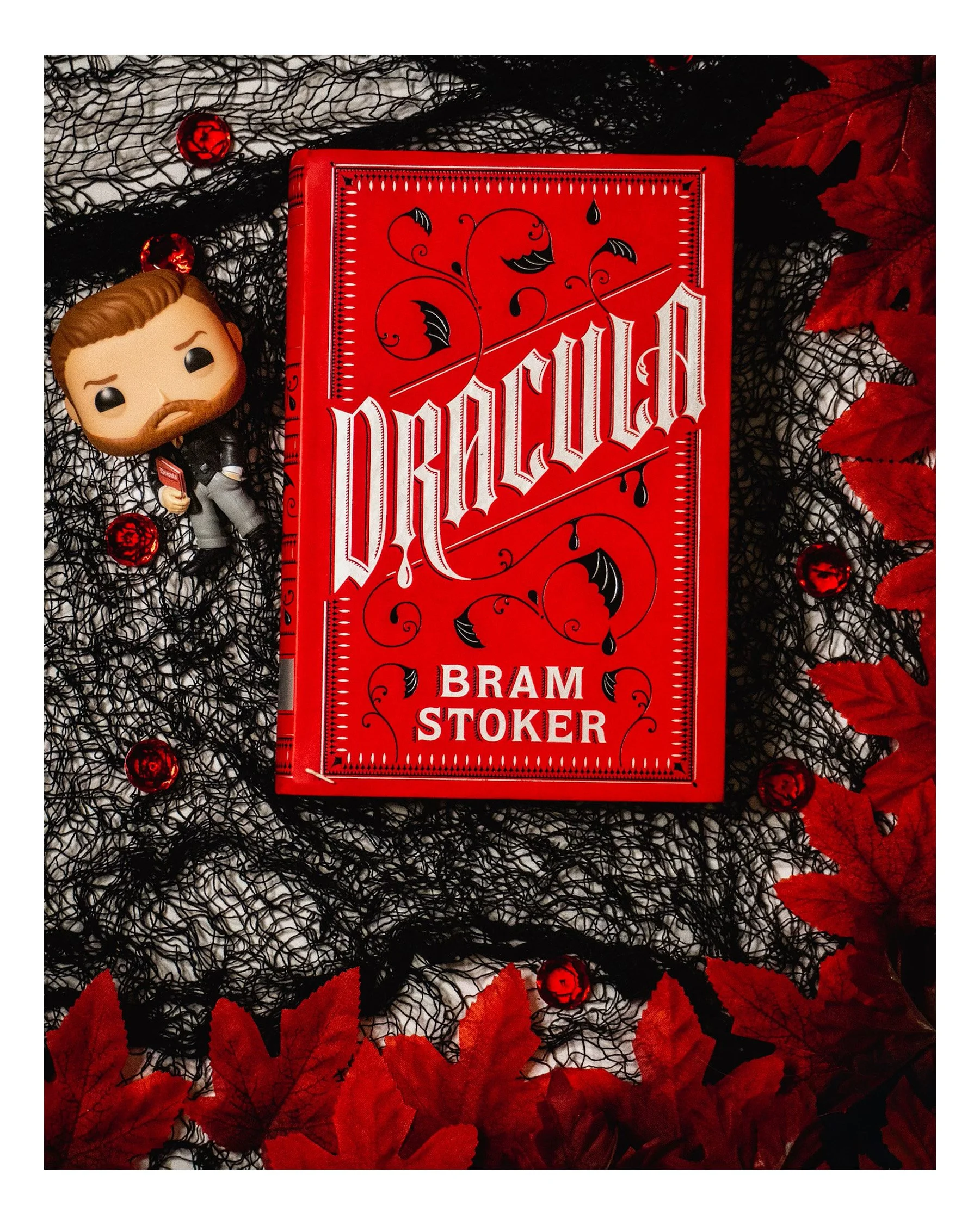 Red book titled 'Futher' by Bram Stoker is placed on black and red decorative leaves with black netting and red decorative ornaments.