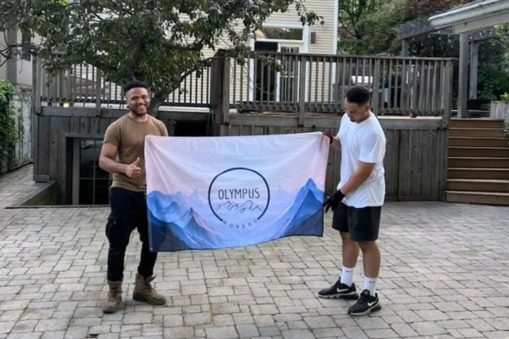 Two men holding an Olympus Movers banner outdoors on a paved surface with a wooden fence and house in the background.