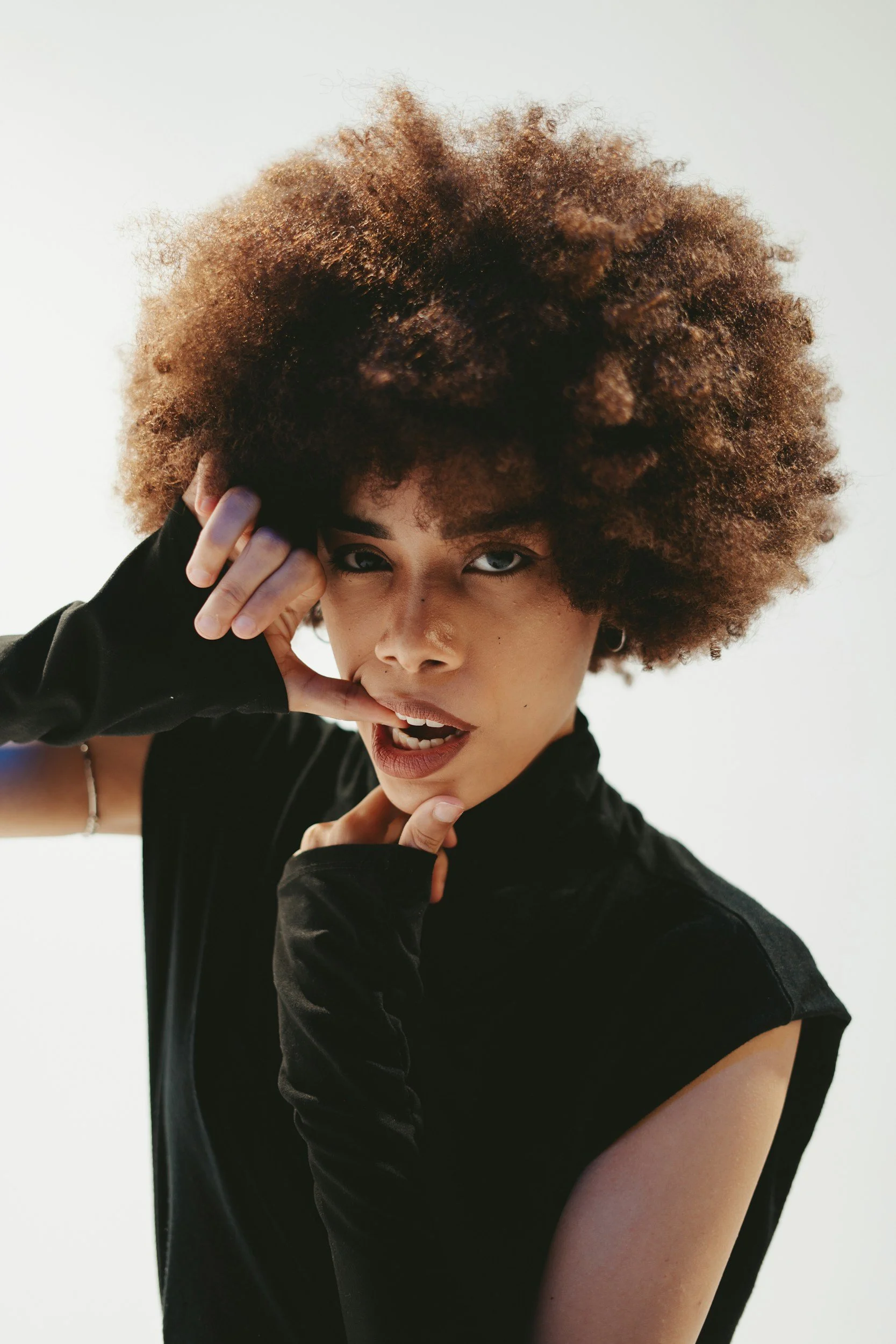 A woman with curly, voluminous hair and piercing blue eyes poses with her hand near her face against a white background, wearing a black top and fingerless gloves.