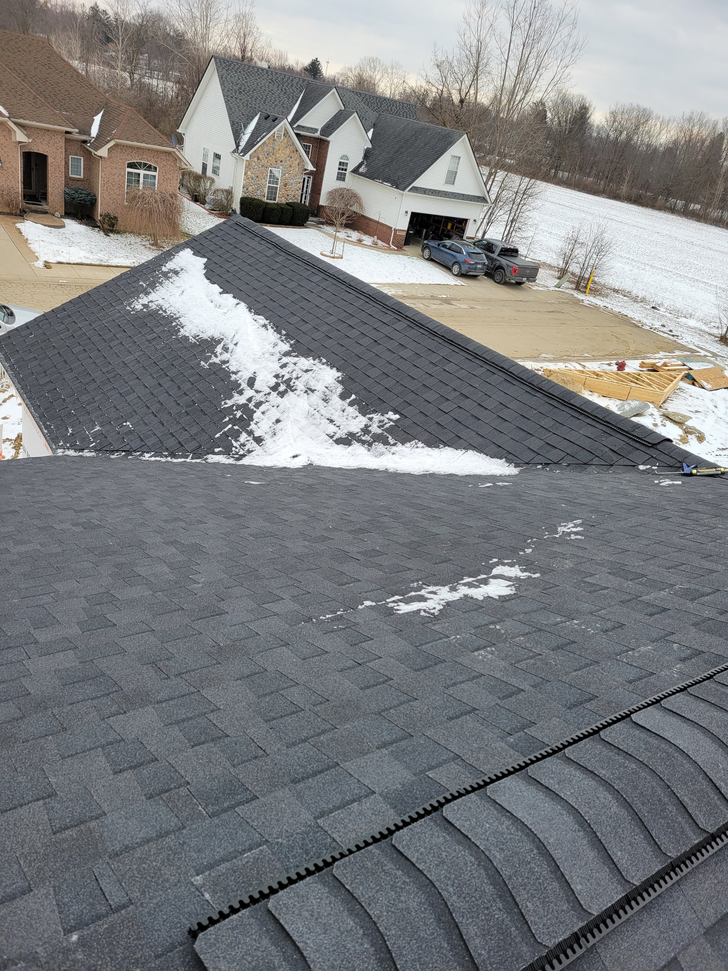 View from a roof showing black shingles with some snow and ice buildup, neighboring houses, a driveway, and an open field with trees in winter.