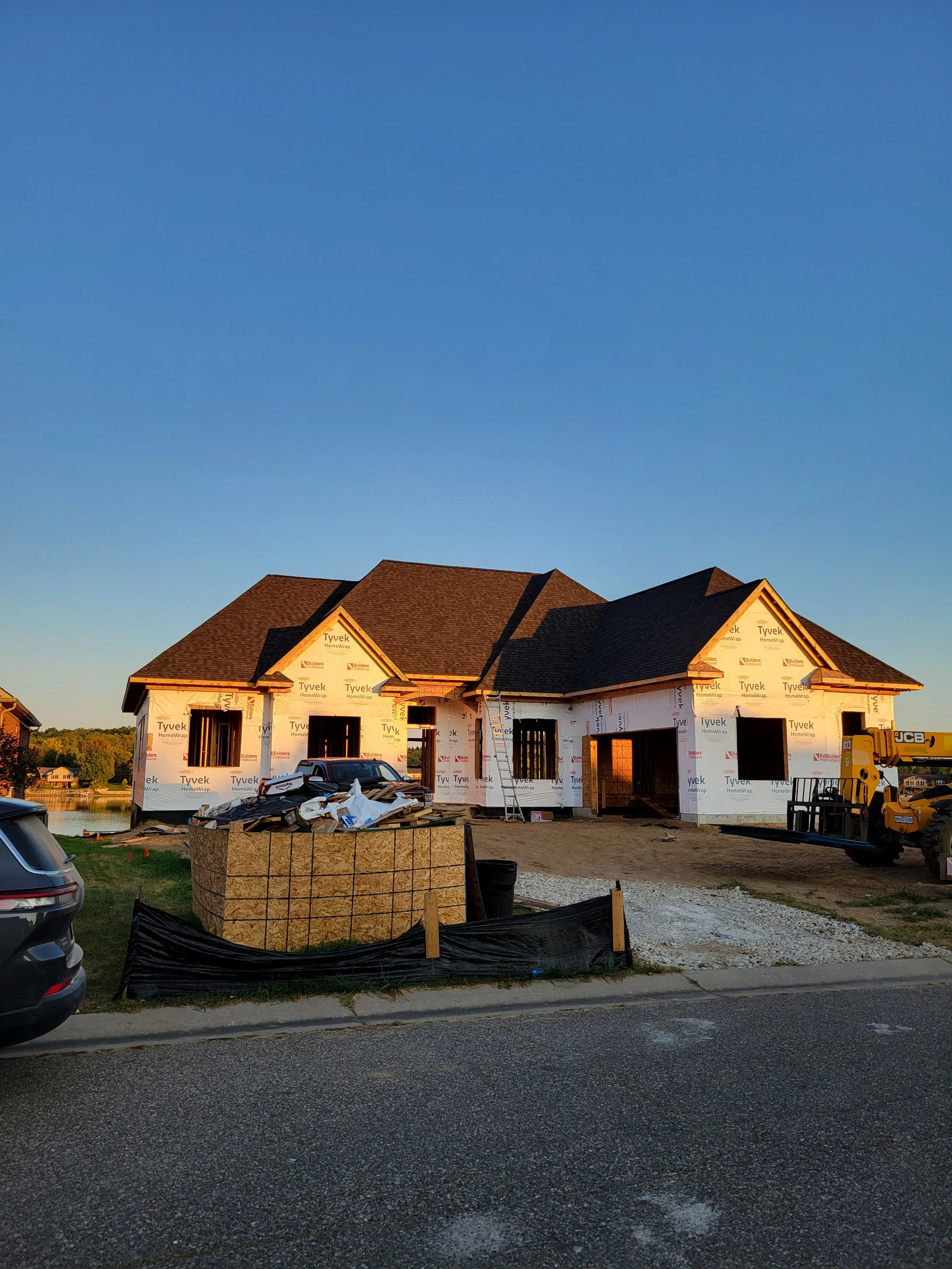A house under construction with a brown roof, partially covered in Tyvek building wrap. Construction equipment and materials are in the front yard, and a small body of water is nearby. Cars are parked on the street in front of the house, and the sky is clear with no clouds.