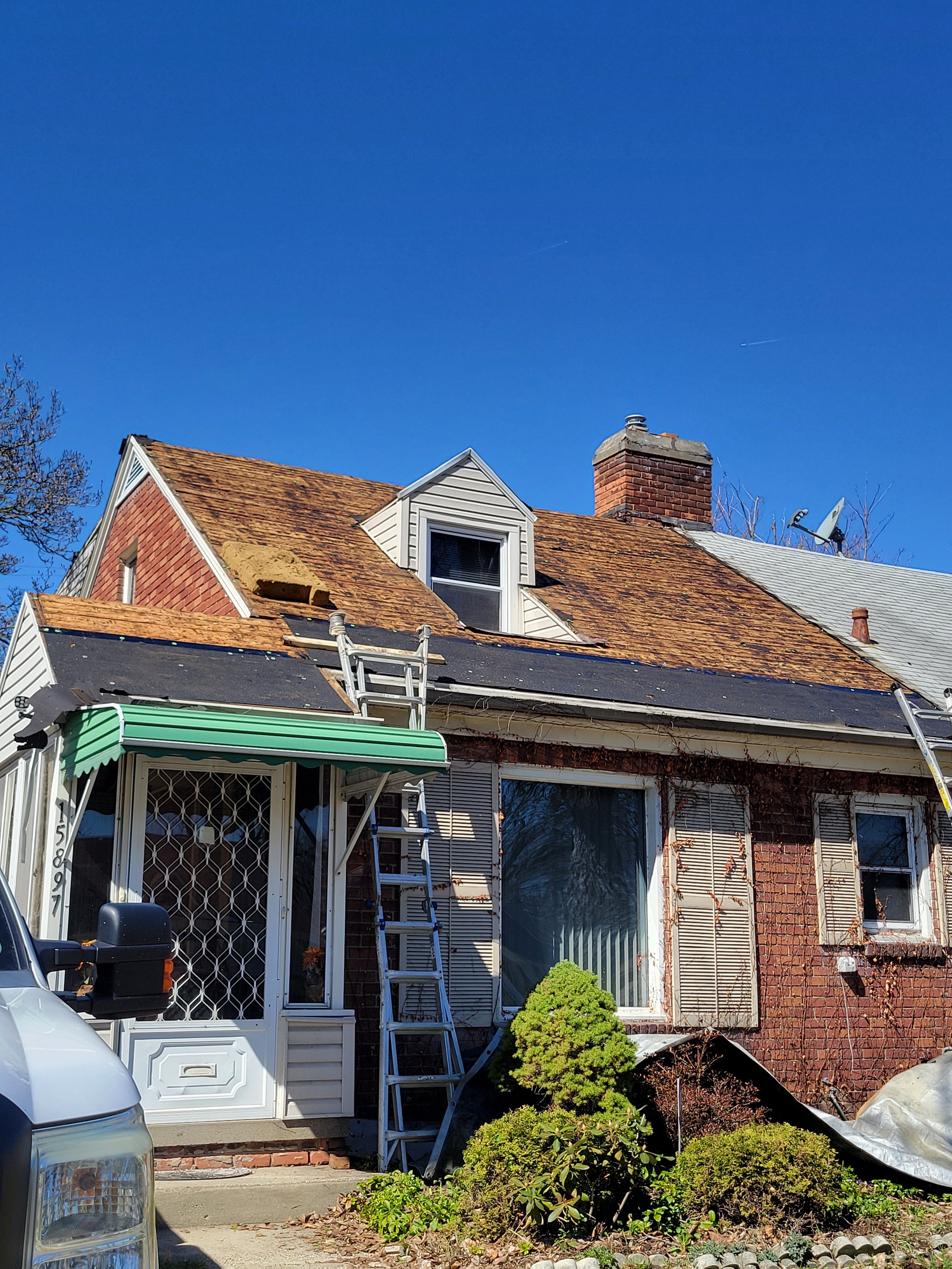House with a brick and white exterior, undergoing roof repairs with ladders and roofing materials. Green bushes in front, clear blue sky above.