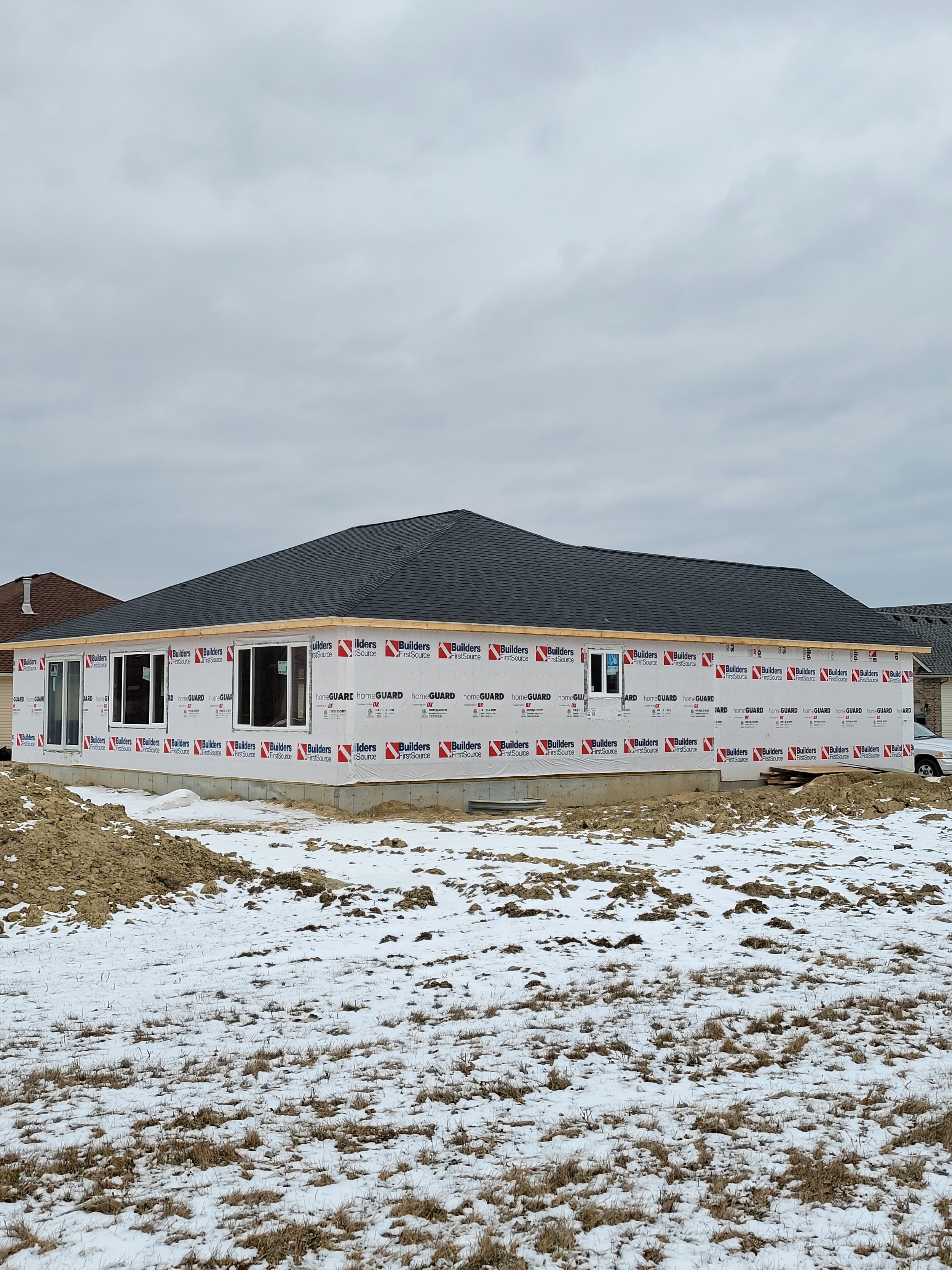 The image shows a house under construction with a black roof and a wrapped exterior. The house is in a snowy, muddy field with patches of snow and dirt. The sky above is cloudy.