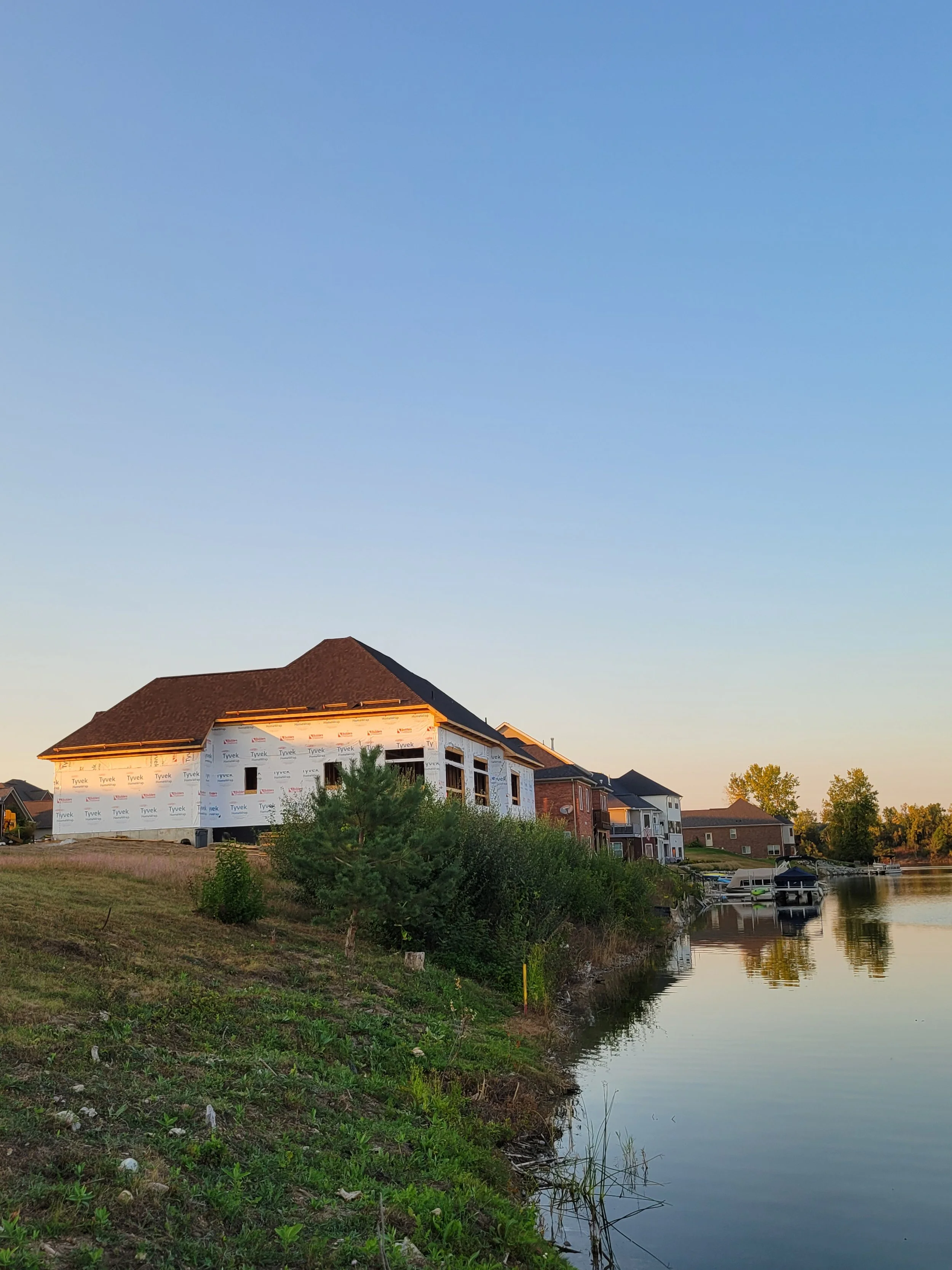 A house under construction with a partially finished exterior, situated next to a body of water with boats docked, and surrounded by greenery, with other completed houses in the background, all under a clear blue sky.