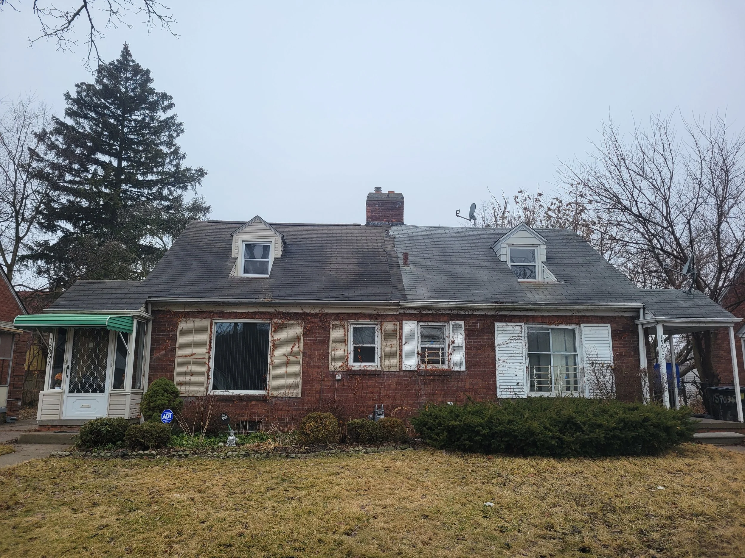 A two-story brick house with a gray shingle roof, two small dormer windows, and a brick chimney. The front yard has shaggy grass, small bushes, and an ADT security sign. Surrounding the house are deciduous and evergreen trees with no leaves, indicating winter.