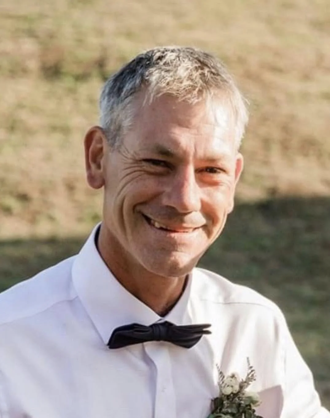 Smiling man with short gray hair wearing a white dress shirt, black bow tie, and boutonniere, outdoors on a sunny day.