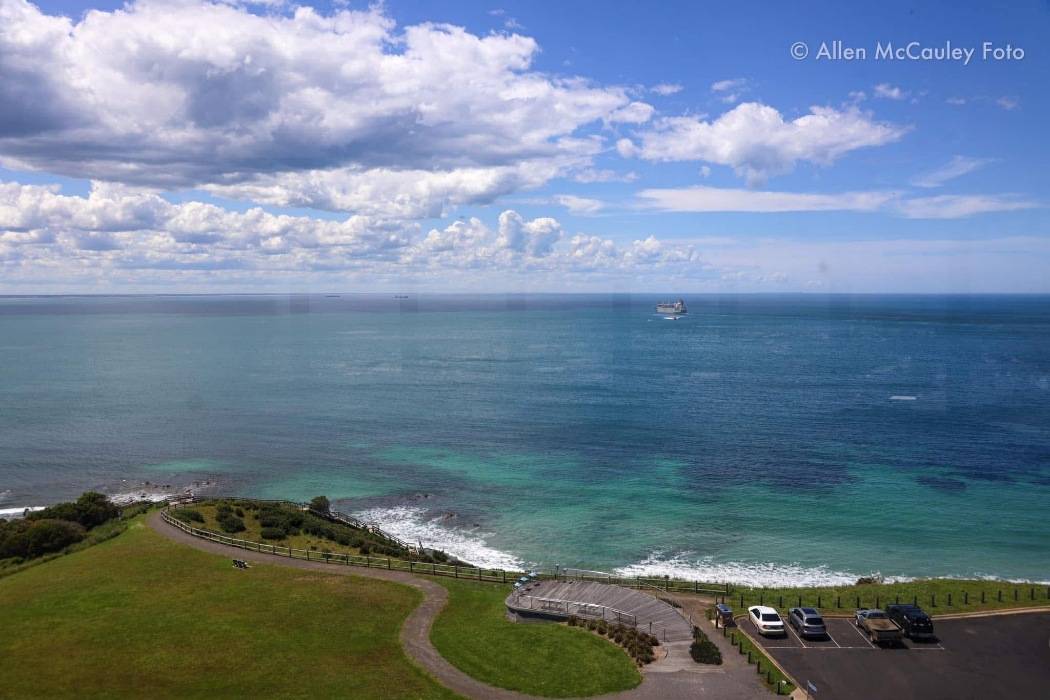 A coastal scene with a grassy park area in the foreground, a pathway, and parked cars. The ocean with a cargo ship and smaller boats is visible, along with a cloudy blue sky.