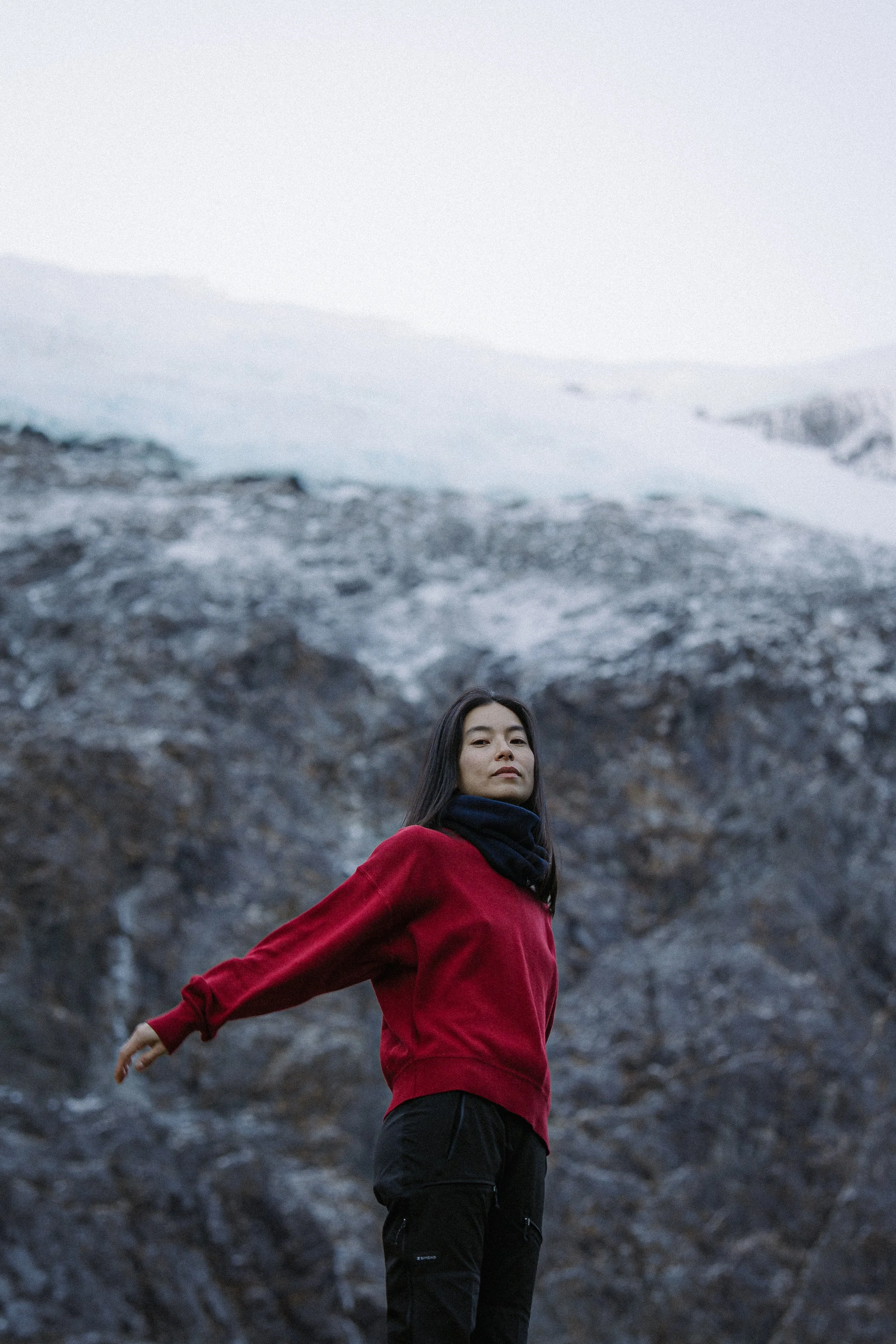 A woman in a red jacket and black pants stands outdoors with rocky mountains and snow in the background. Her arms are extended to the sides, and she looks confidently at the camera.