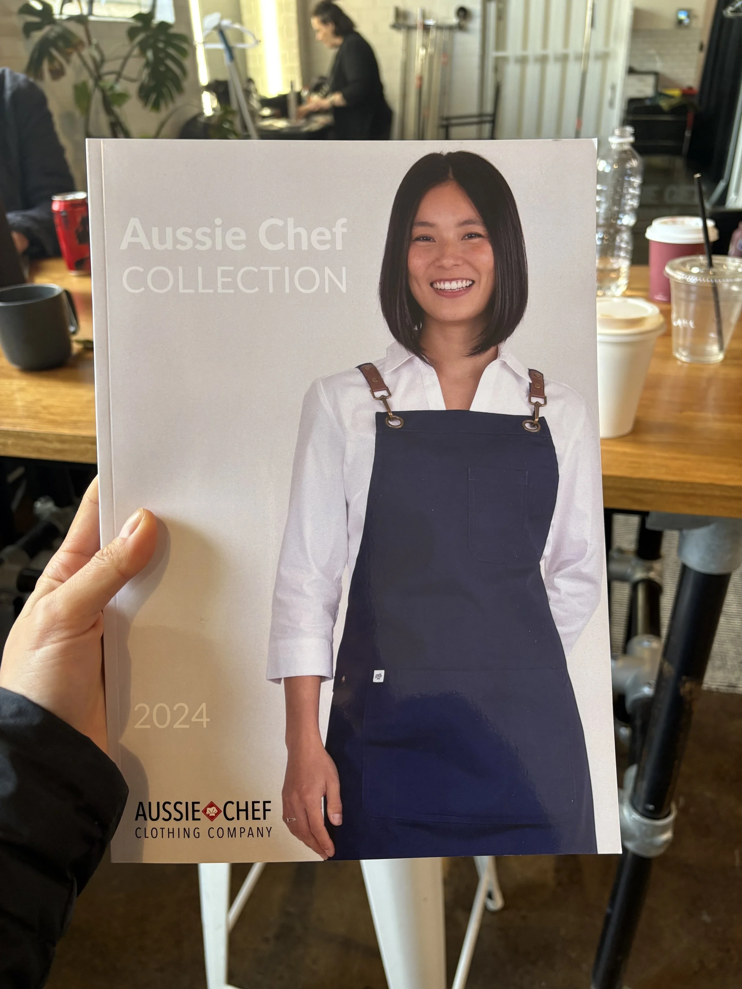 A promotional flyer for Aussie Chef clothing company featuring a smiling woman with shoulder-length black hair, wearing a white shirt and a dark apron, on a casual dining table background.