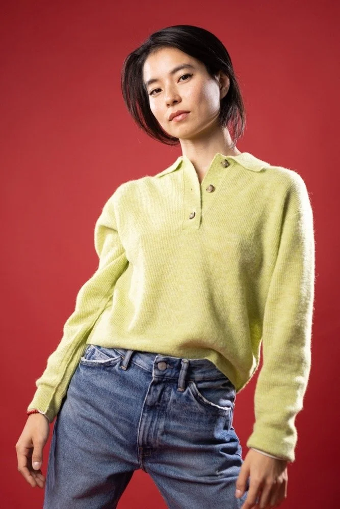 A woman with short black hair wearing a yellow sweater and blue jeans against a red background.