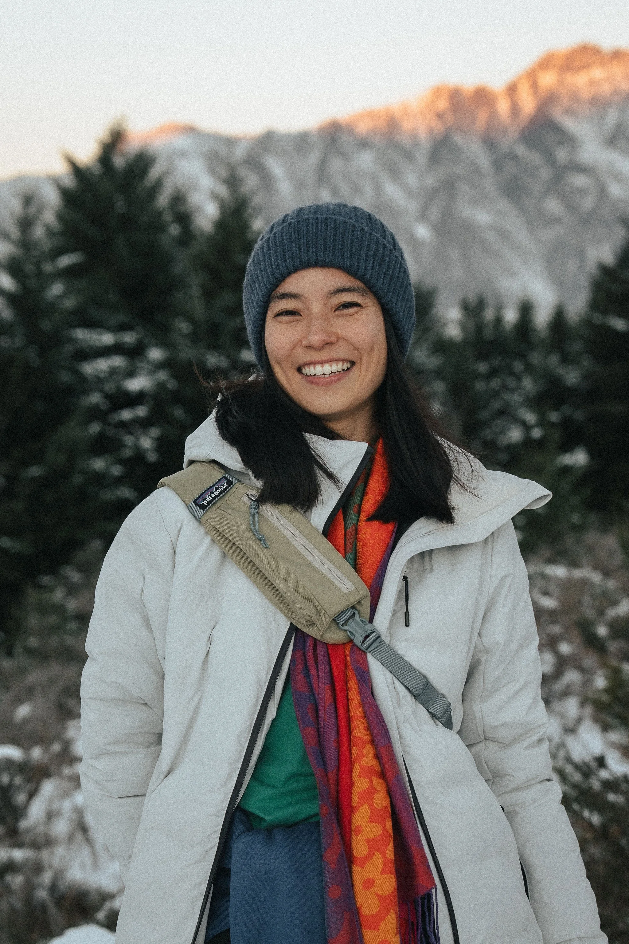 Woman smiling outdoors in a snowy mountainous landscape, wearing a blue beanie, white jacket, and colorful scarf.