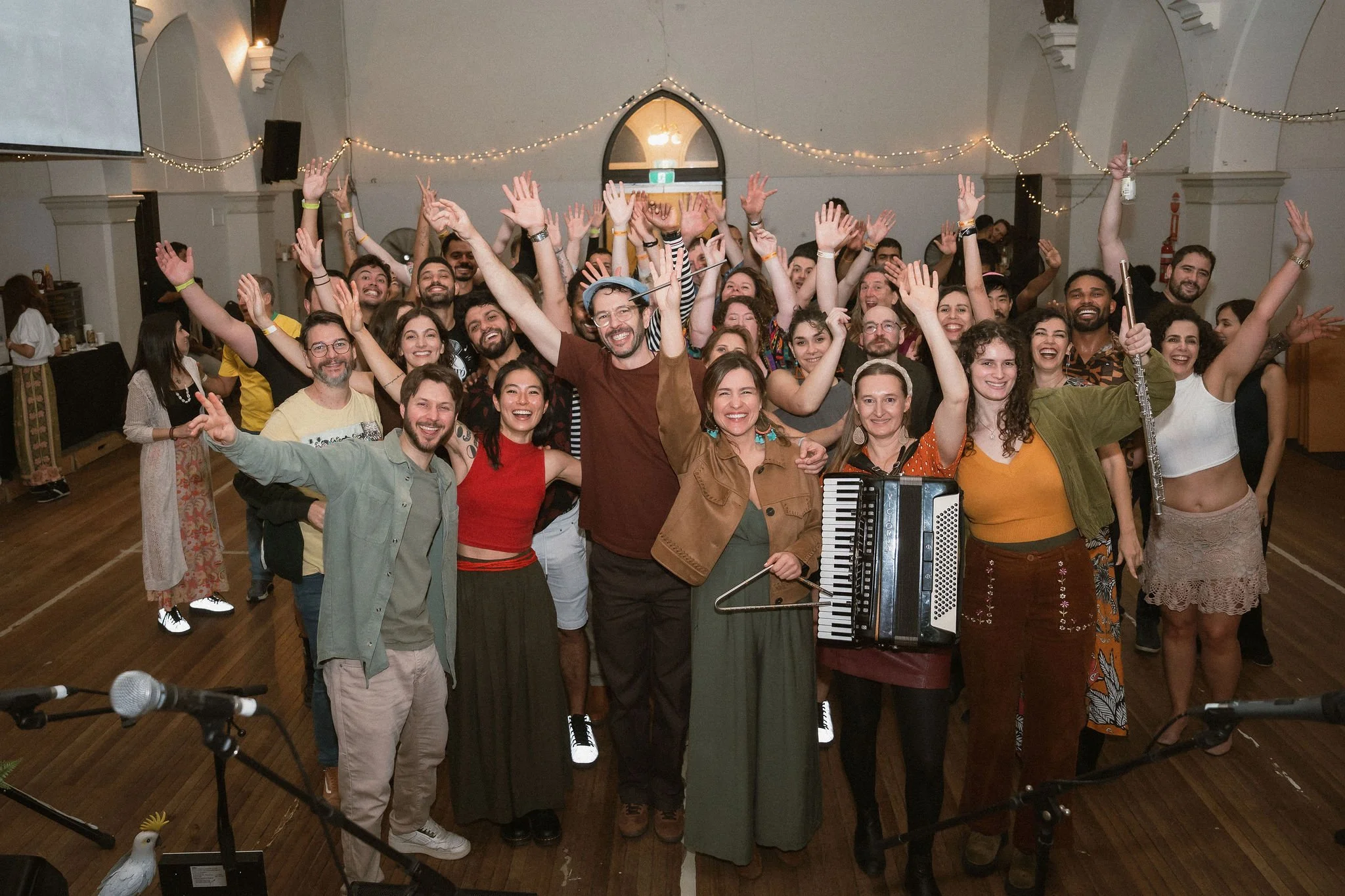 A large group of people gathered in a hall, celebrating and smiling for the photo. Some are raising their hands, and someone in the front is holding an accordion. The hall is decorated with string lights.
