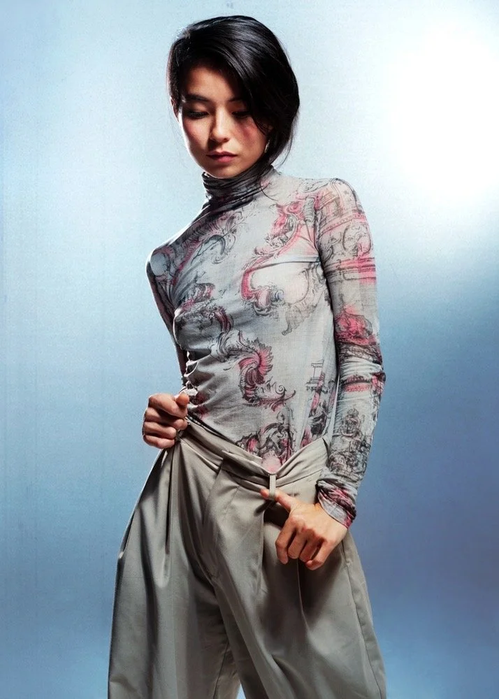 A young woman with short dark hair wearing a fitted long-sleeve shirt with a pink and gray abstract pattern and loose beige trousers, standing against a light blue background.