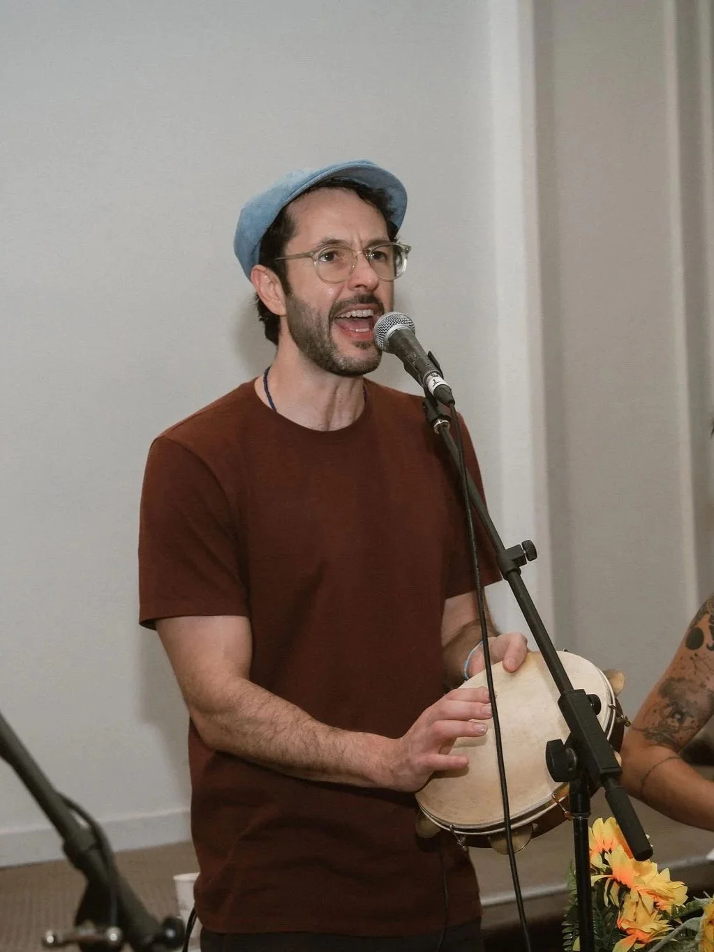 A man singing into a microphone while playing a tambourine. He wears glasses, a blue hat, and a maroon T-shirt.