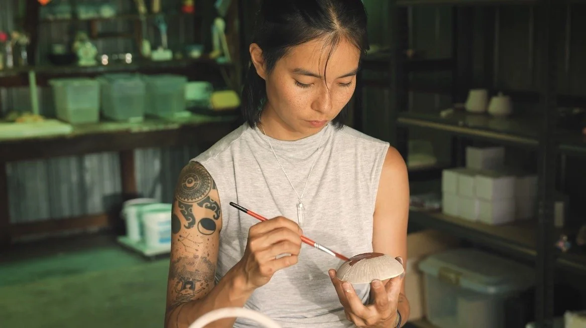 A woman with tattoos wearing a gray sleeveless shirt is painting a ceramic object with a small brush in a workshop.