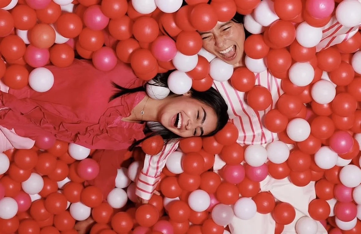 Two women are having fun lying in a ball pit filled with red, white, and pink plastic balls. One woman is wearing a red dress, and the other is wearing a striped shirt. They are both smiling and appear to be enjoying themselves.