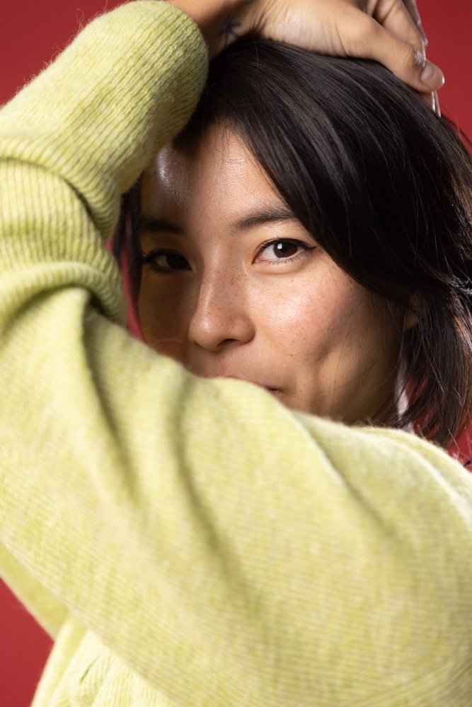Close-up of a young woman with dark hair, wearing a yellow sweater, resting her arm on her head and looking at the camera with a neutral expression.