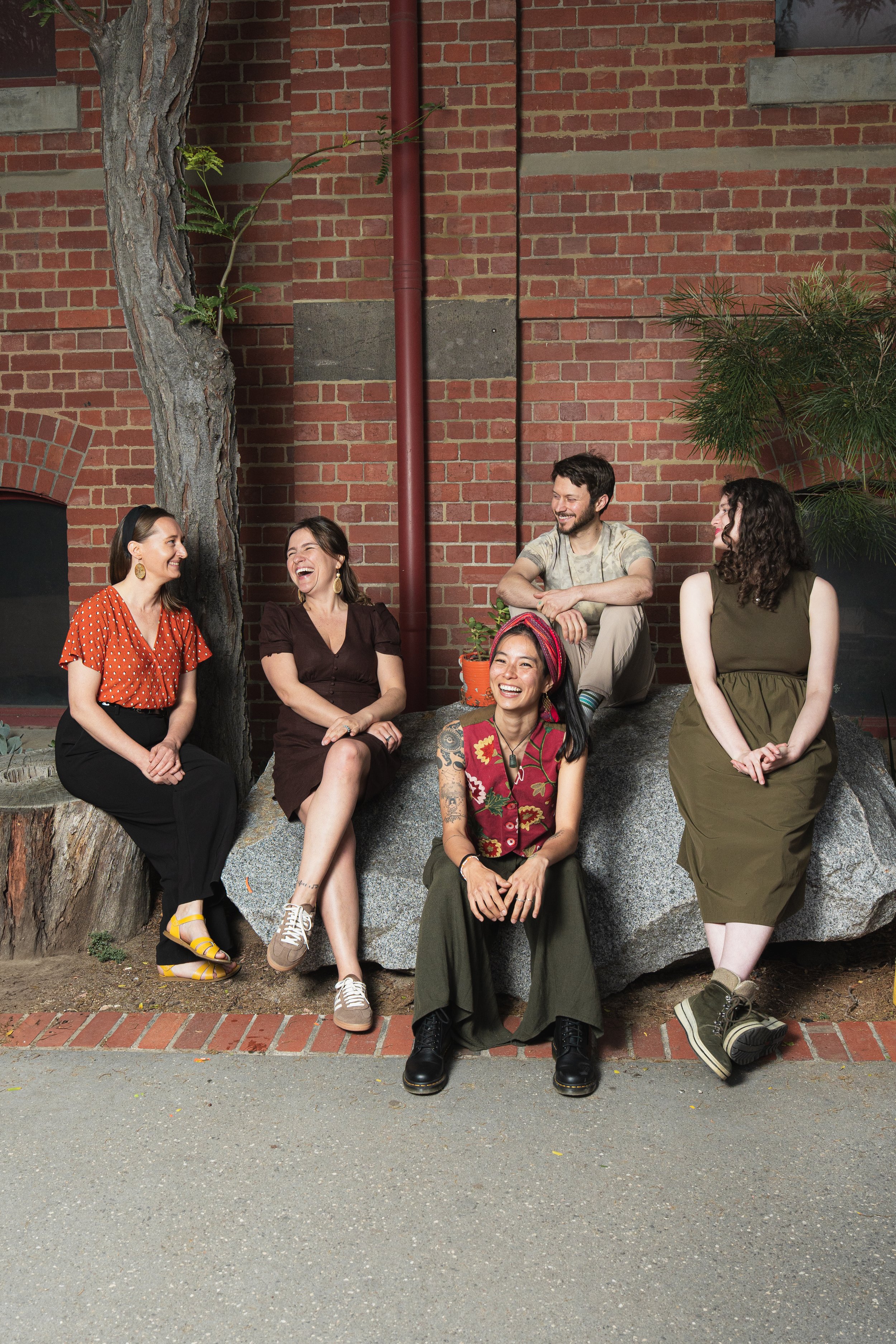 A group of six diverse people sitting and standing on a large rock, laughing and talking, against a brick wall with some greenery nearby.
