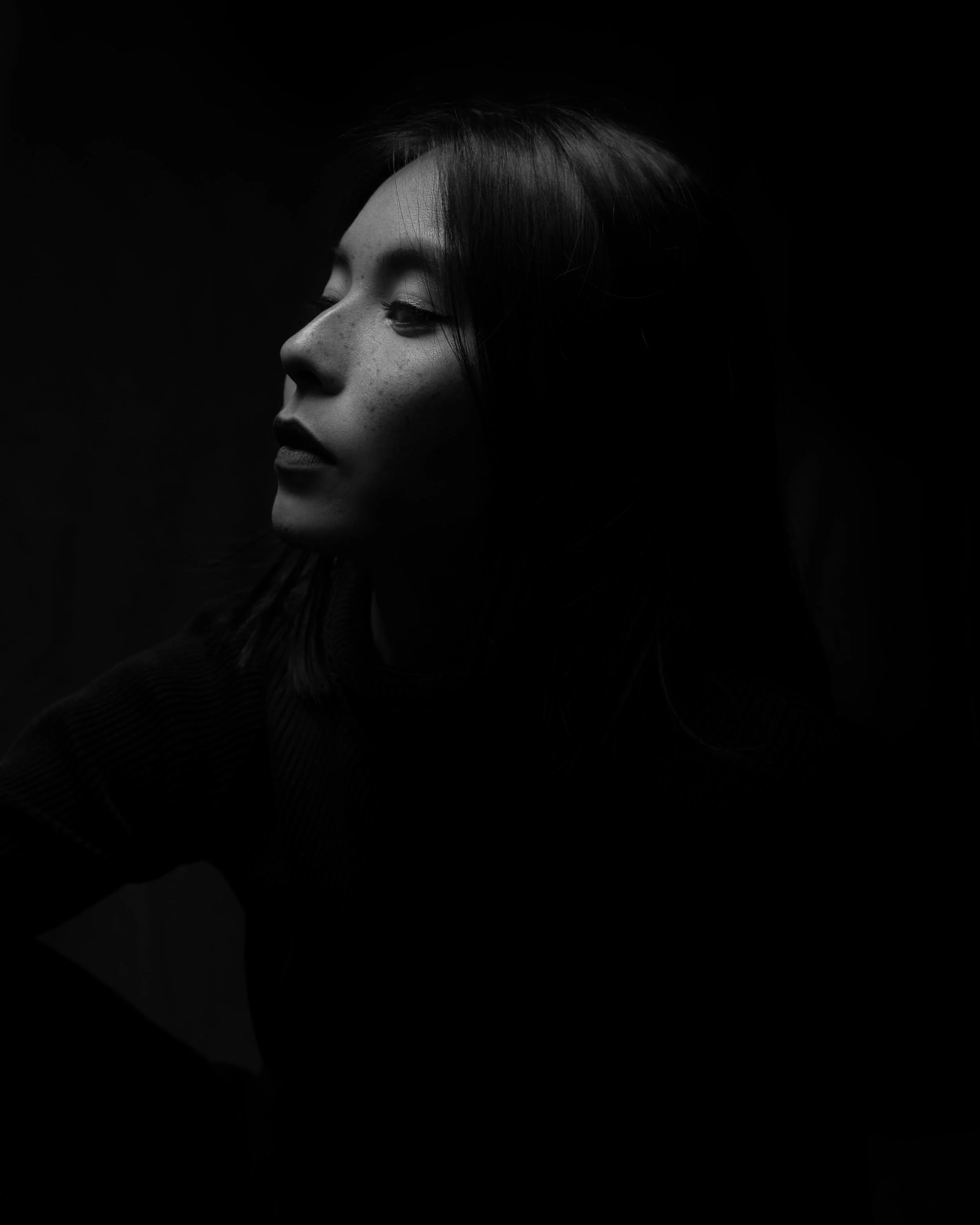 A person with closed eyes and long dark hair, facing to the left, against a black background.