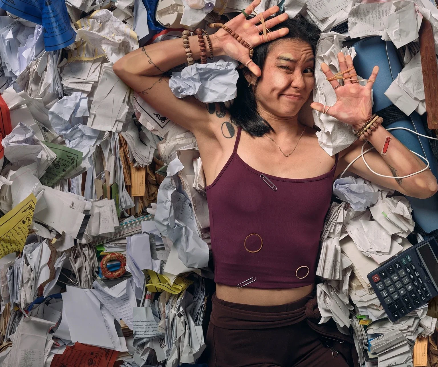 A woman with tattoos and multiple bracelets lying among crumpled papers, using a calculator and making a playful face with one eye closed and hands near her face.
