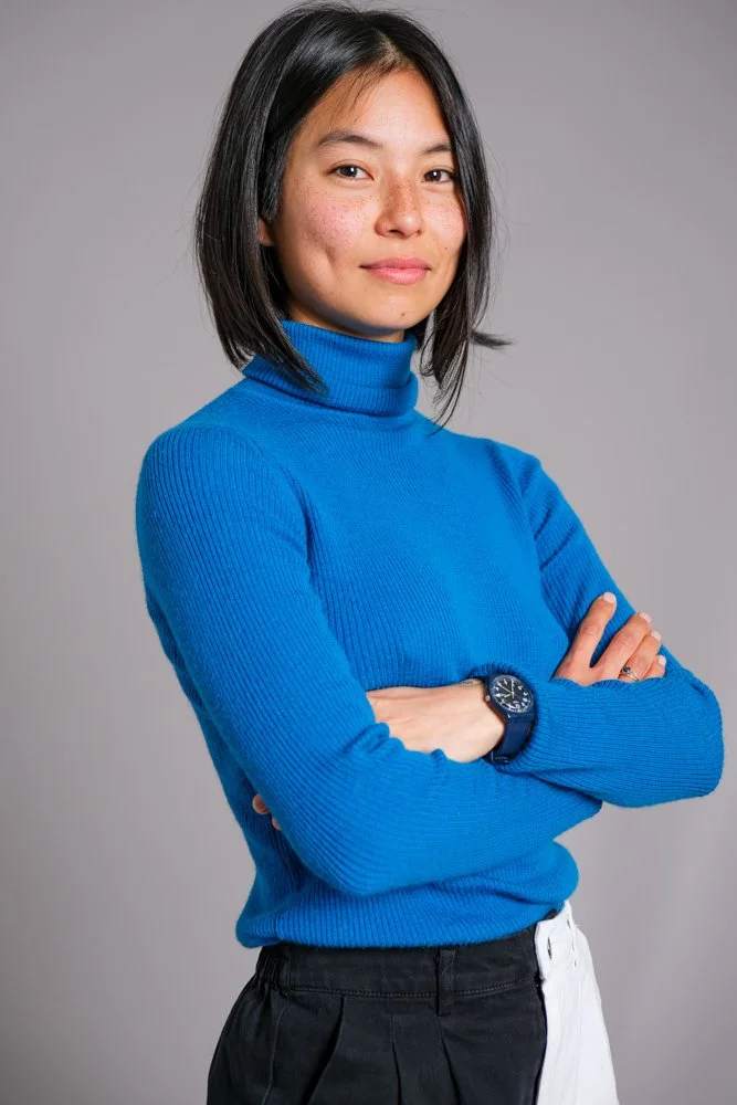 A young woman with black hair, wearing a blue turtleneck sweater and a wristwatch, standing with her arms crossed against a plain gray background.
