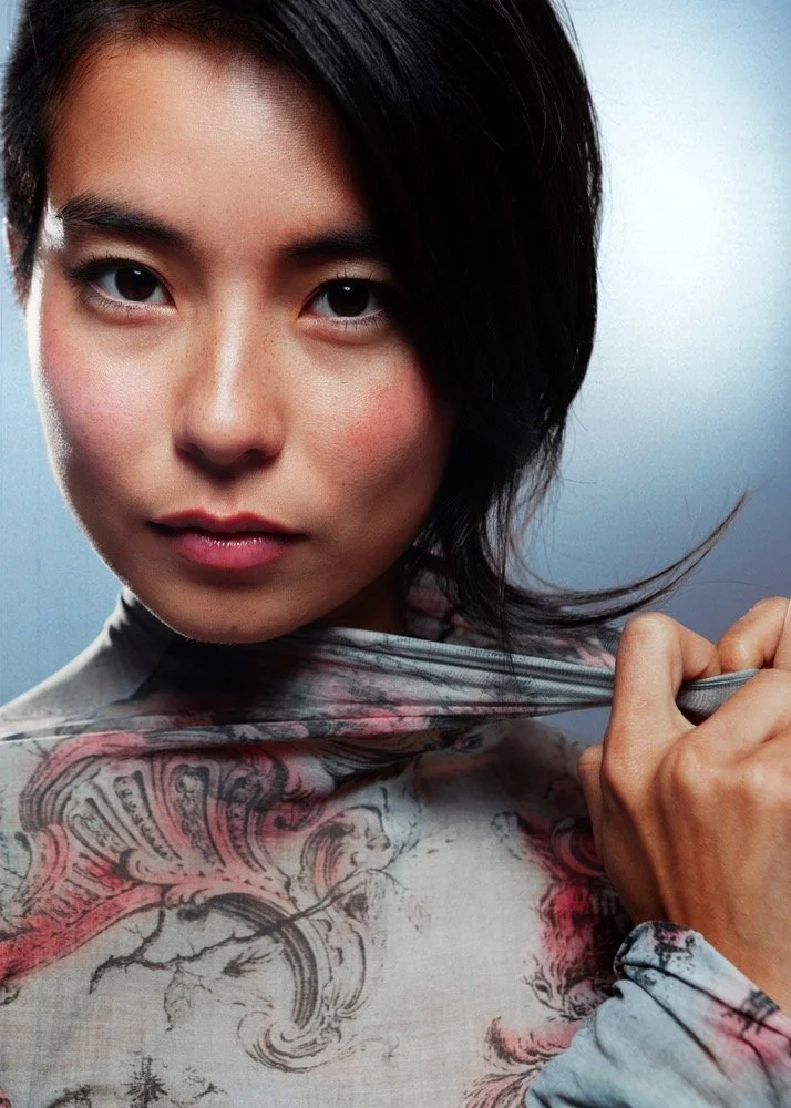 A woman with short black hair holding the collar of her patterned shirt, looking directly at the camera with a neutral expression.