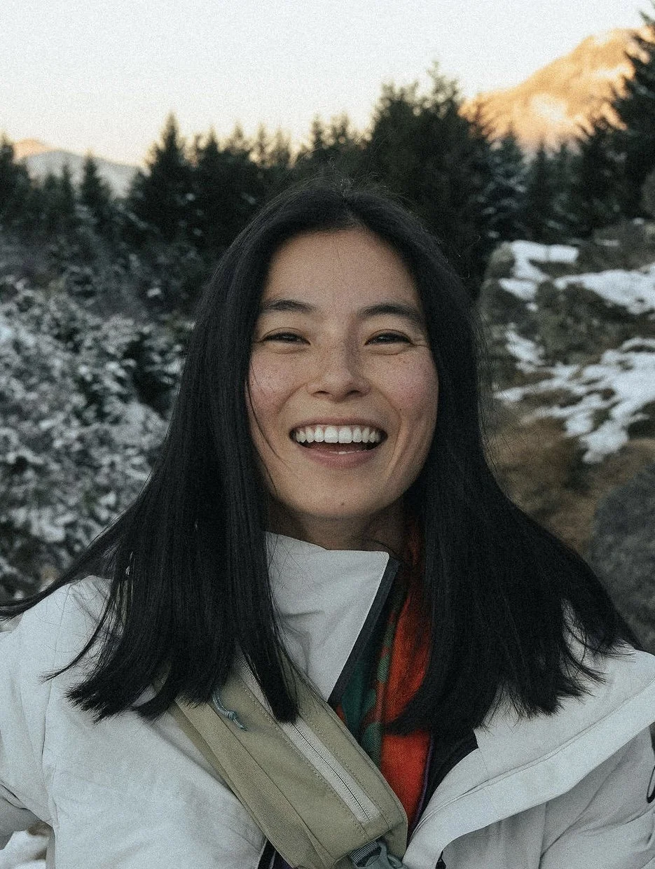 A woman smiling outdoors in a snowy, mountainous landscape with pine trees and peaks in the background.