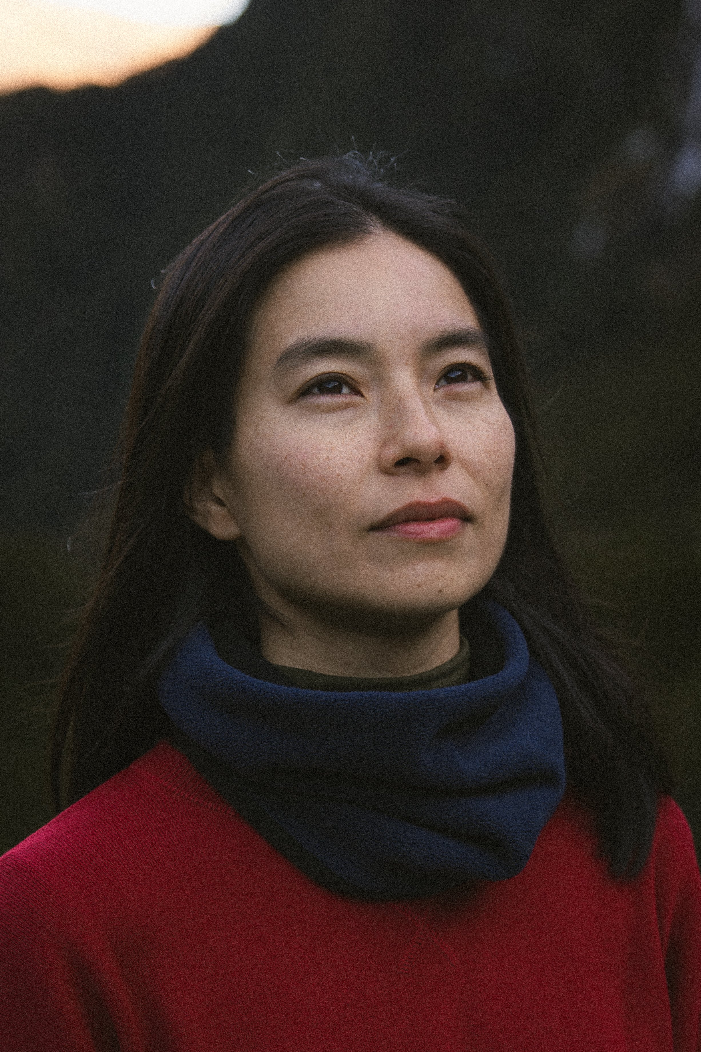 Close-up of a young woman with dark hair, wearing a red and blue jacket, outdoors during sunset, with a dark silhouette of a mountain in the background.