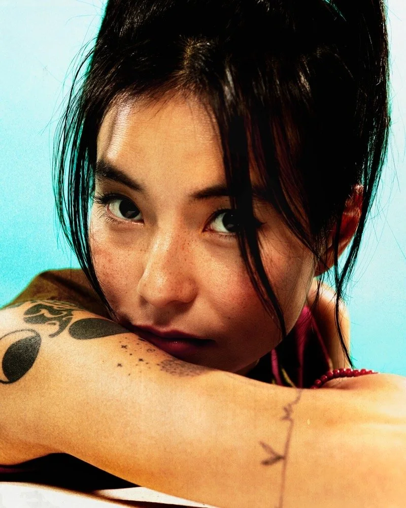 Close-up portrait of a woman with black hair, tattoos on her arm, and intense gaze, resting her arm on a surface against a light blue background.