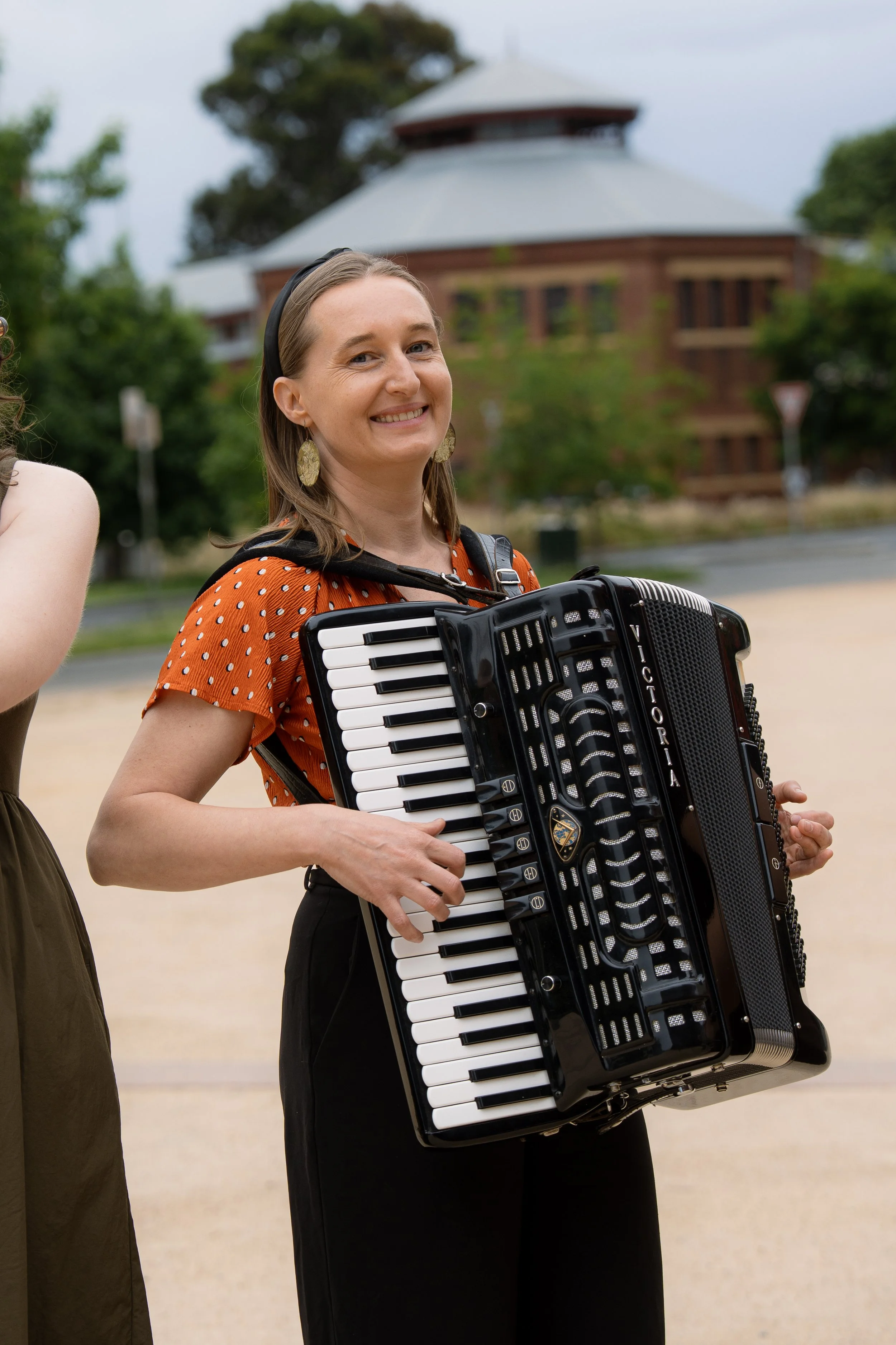 A smiling woman holding a black and white accordion outdoors with a large round building and trees in the background.
