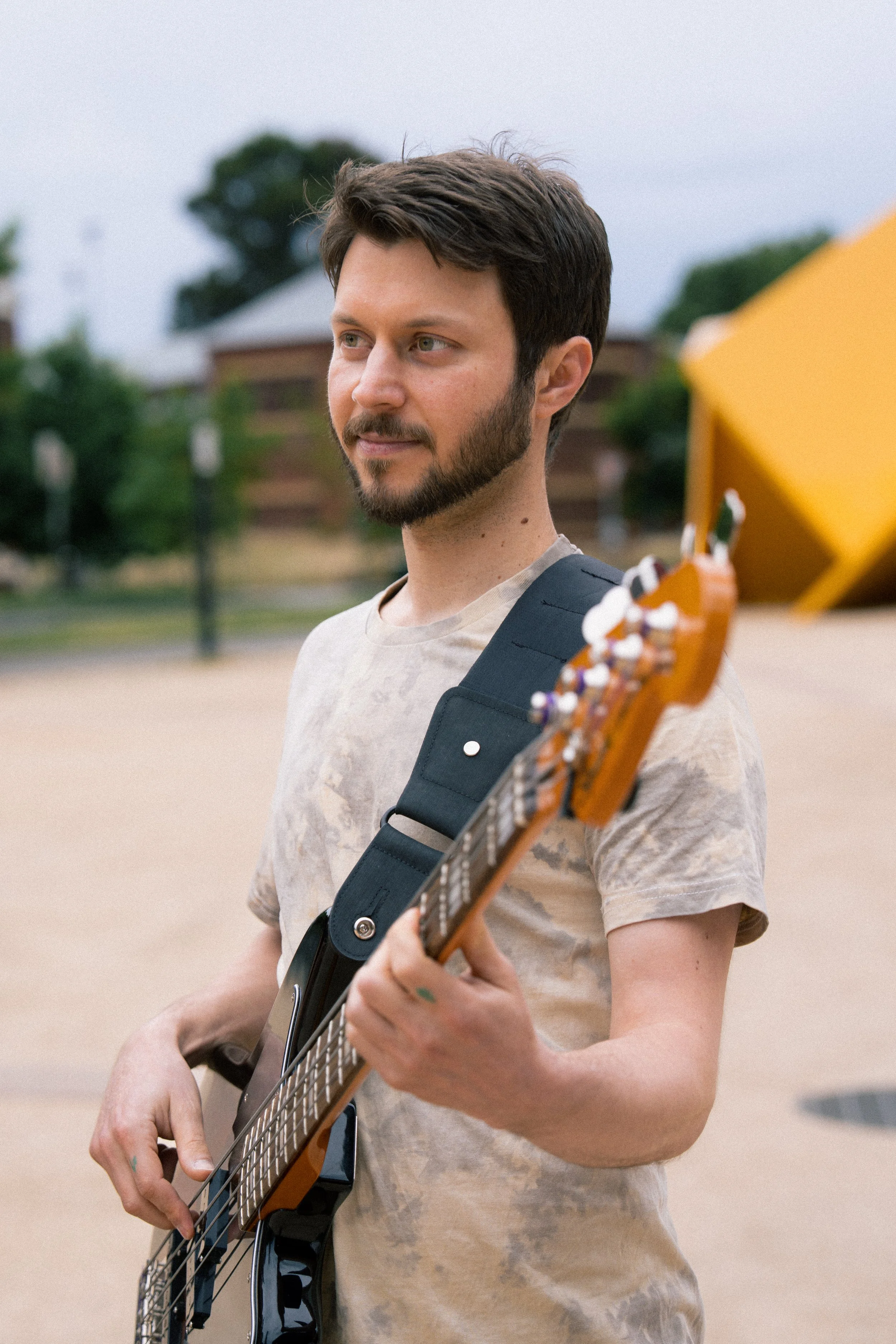 A young man with brown hair and a beard, wearing a light-colored t-shirt, playing an electric guitar outdoors. The background shows trees and a building, with a yellow umbrella partially visible on the right.