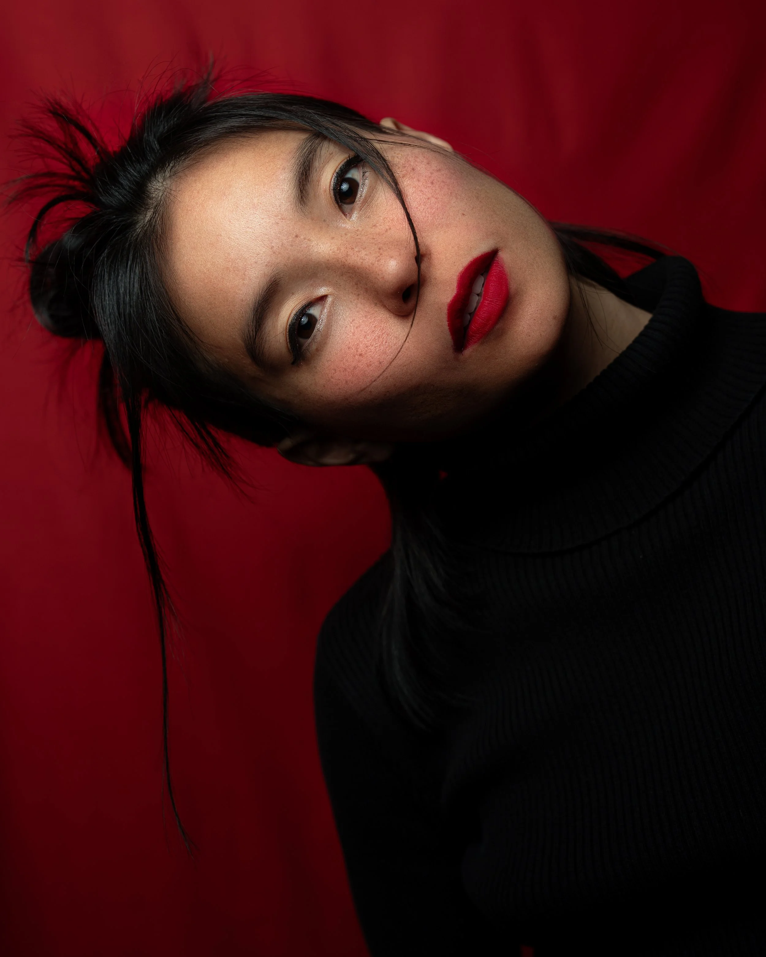 Close-up of woman's face with black hair, red lipstick, and a black top, against a red background.