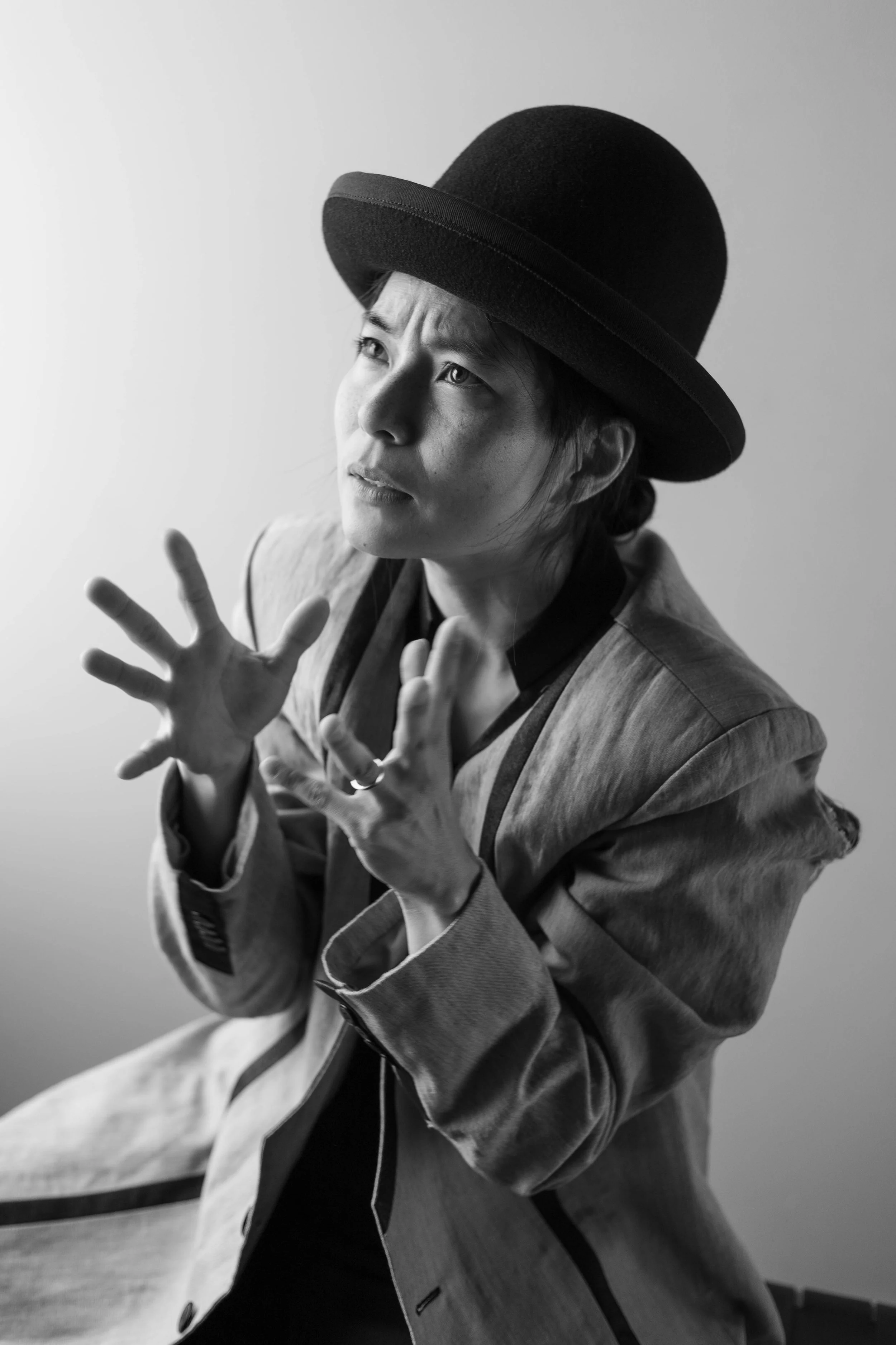 A person wearing a large, wide-brimmed hat, looking upwards with a focused expression, and their hands raised in front of them.