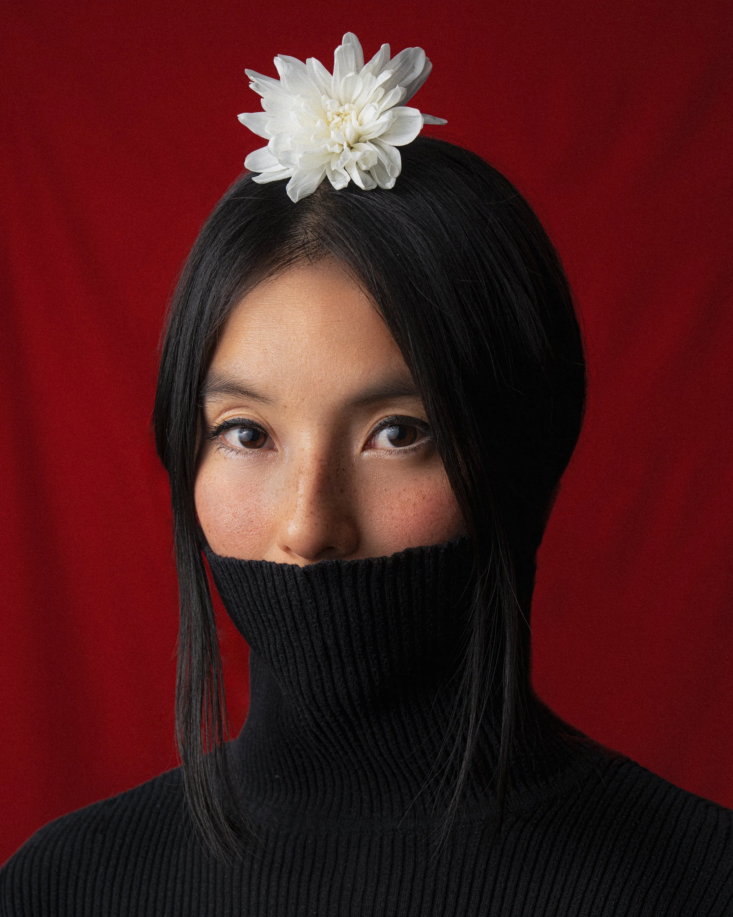 A woman with black hair and blue eyes wearing a black turtleneck sweater and a white flower on her head, set against a red background.