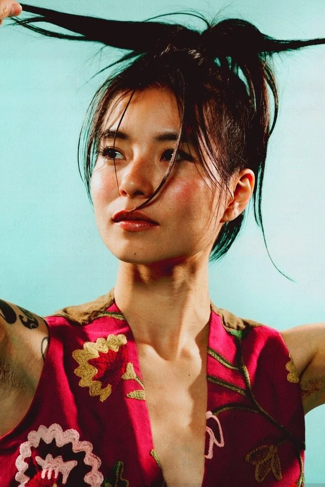A woman with short dark hair styled in an edgy, windswept look, wearing a colorful embroidered top with floral patterns, against a plain turquoise background.