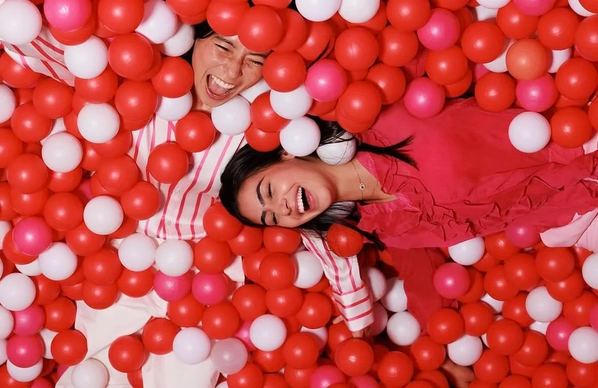 Two women laughing and playing in a ball pit filled with red, white, and pink plastic balls.