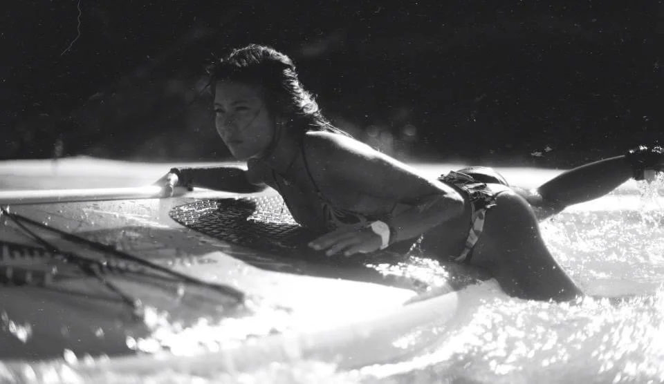 A woman is lying on a surfboard in the water, smiling, with water splashing around her.