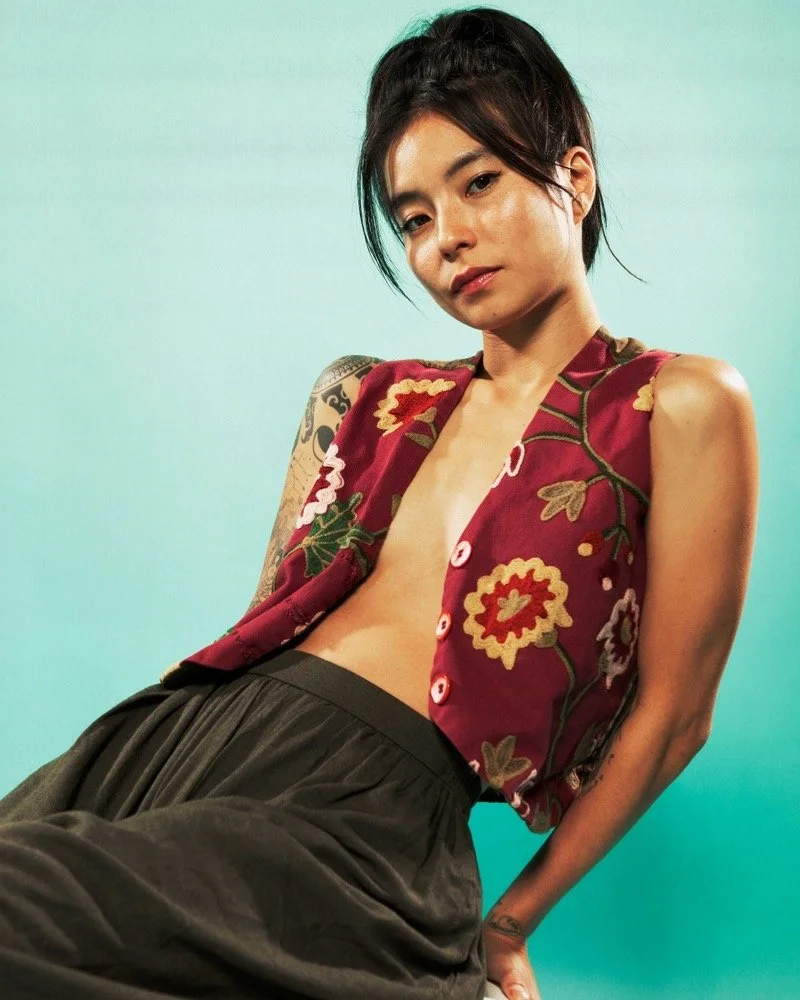 A woman with short black hair and a tattoo on her arm, wearing a sleeveless embroidered vest and dark pants, posing against a gradient blue-green background.