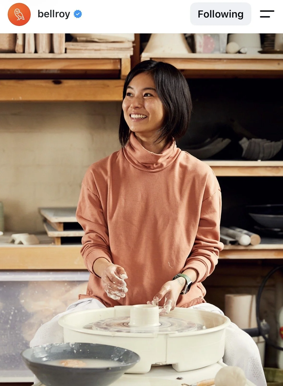 A woman in a peach turtleneck sweater is working on a pottery wheel, shaping clay into a vessel in a ceramics studio. She is smiling and looking to her right.