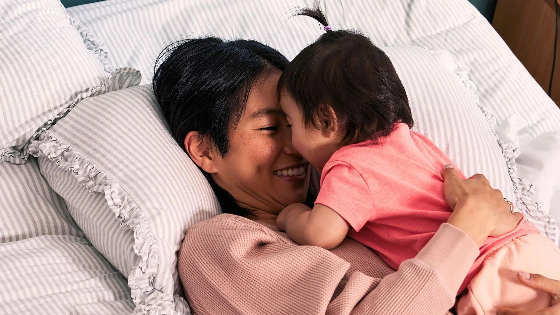 A woman lying on a bed with striped pillows, smiling as she interacts closely with a young girl, who is leaning in to touch foreheads and share a moment.