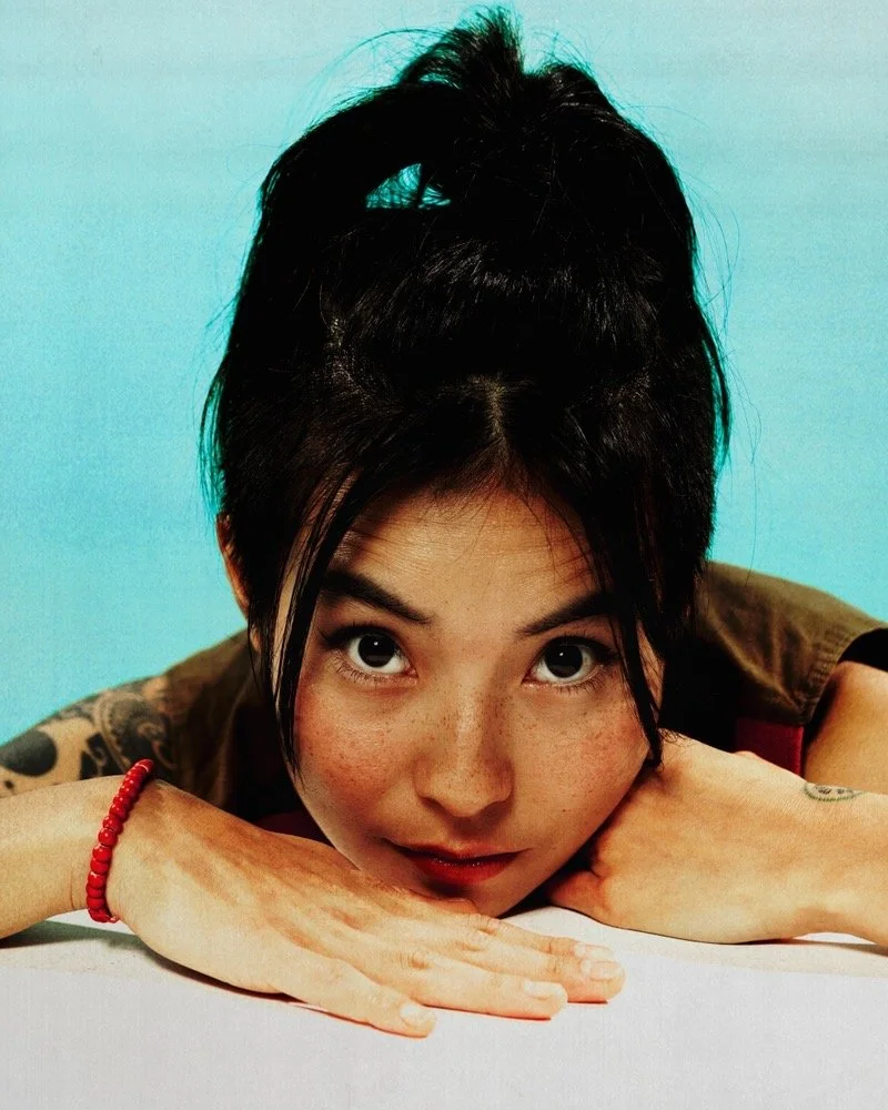 A woman with dark hair styled in a messy bun, lying on her stomach, resting her head and arms on a surface, looking directly at the camera with wide eyes, against a light blue background.