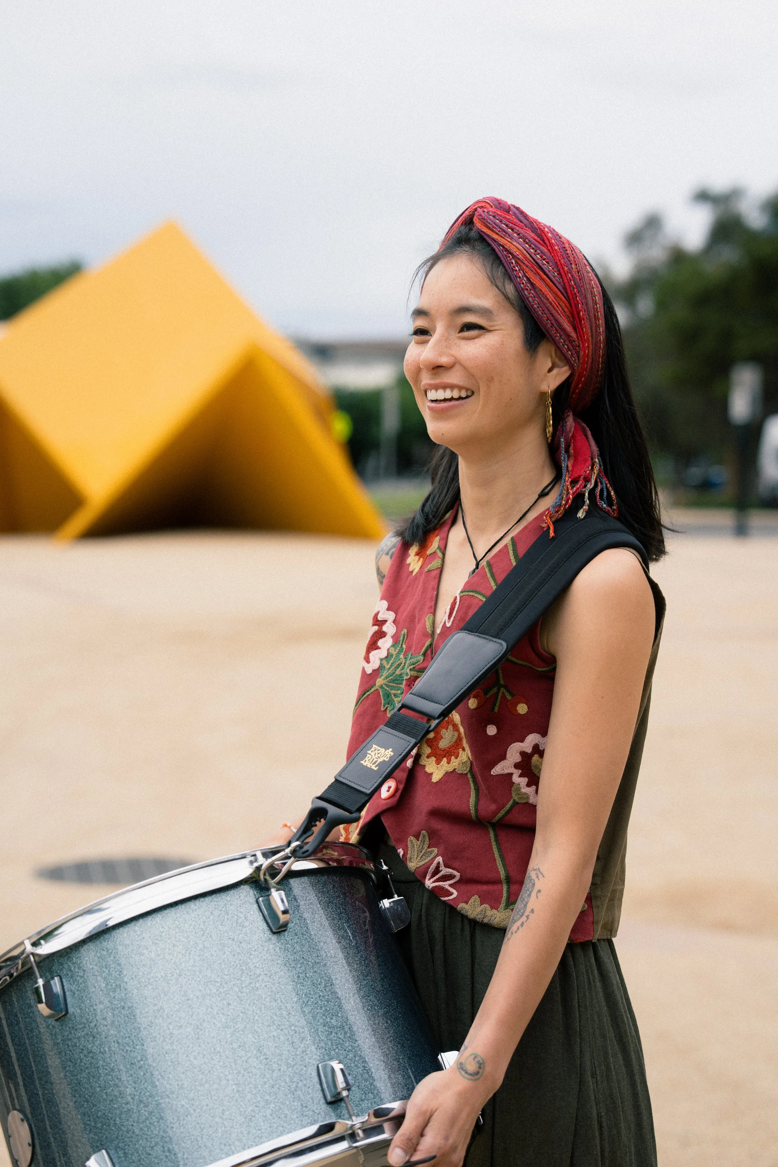 A woman smiling while playing a drum outdoors, wearing a colorful headscarf, earrings, a necklace, and a sleeveless top with embroidery.